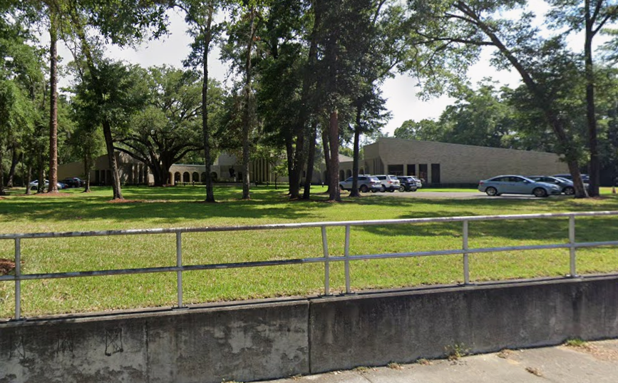 A parking lot with several cars parked in front of a modern, beige-colored building surrounded by green trees, viewed through a metal fence.