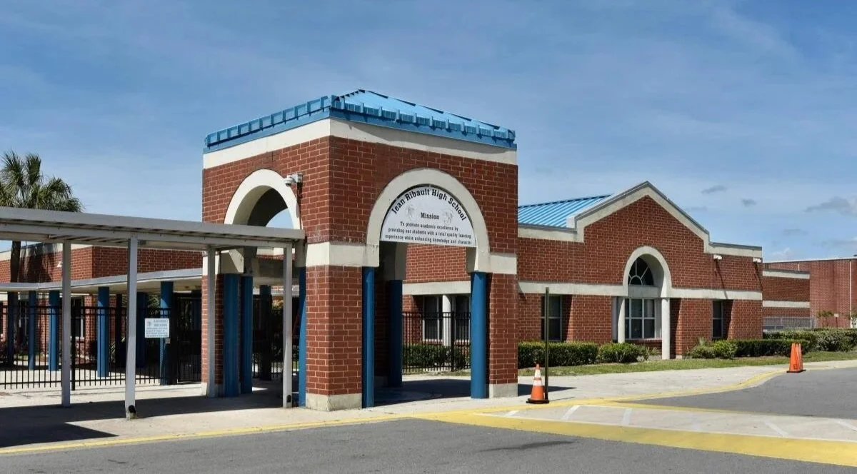 Brick high school building with blue roofing, arched entrance, and security gates, identified as Team Ribbaut High School.