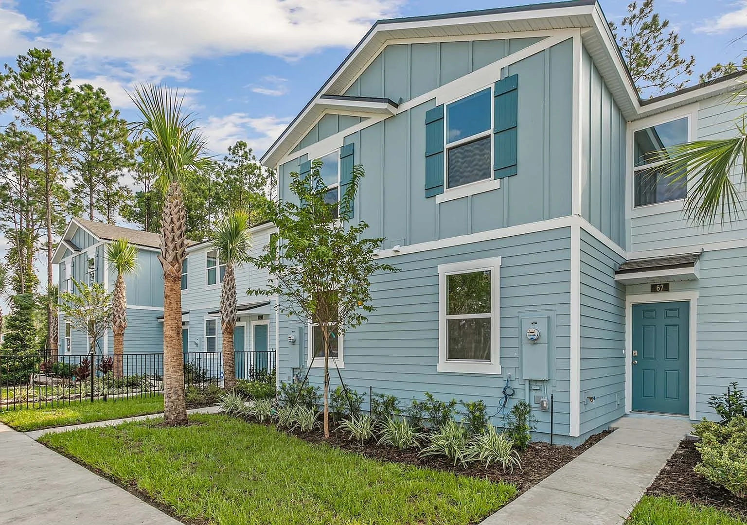 Light blue multi-story townhouses with white trim, surrounded by green lawn, small trees, and palm trees, under a partly cloudy sky.