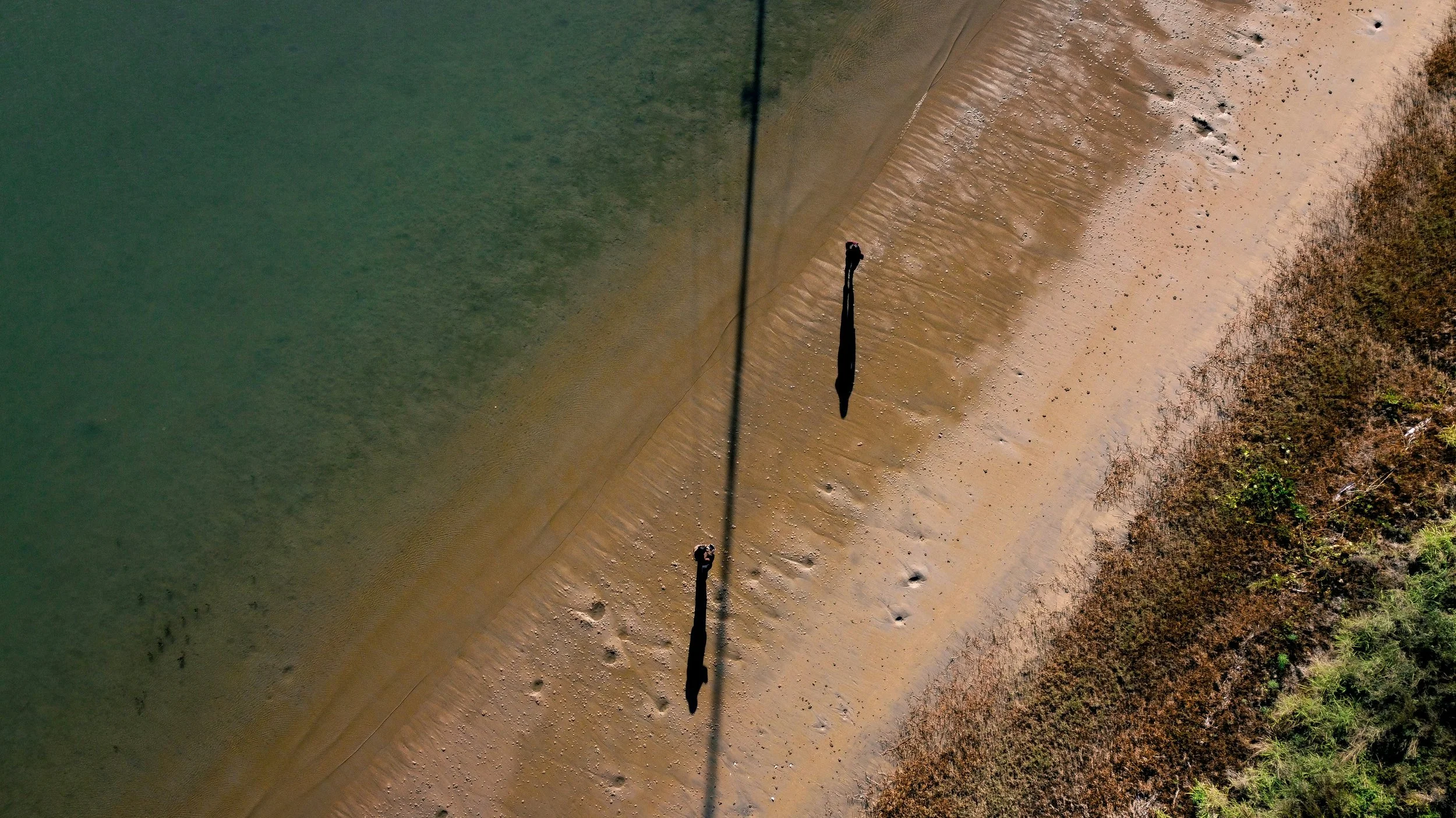 Aerial view of people walking along a sandy beach with footprints, next to green water, and some bushes on the right side.