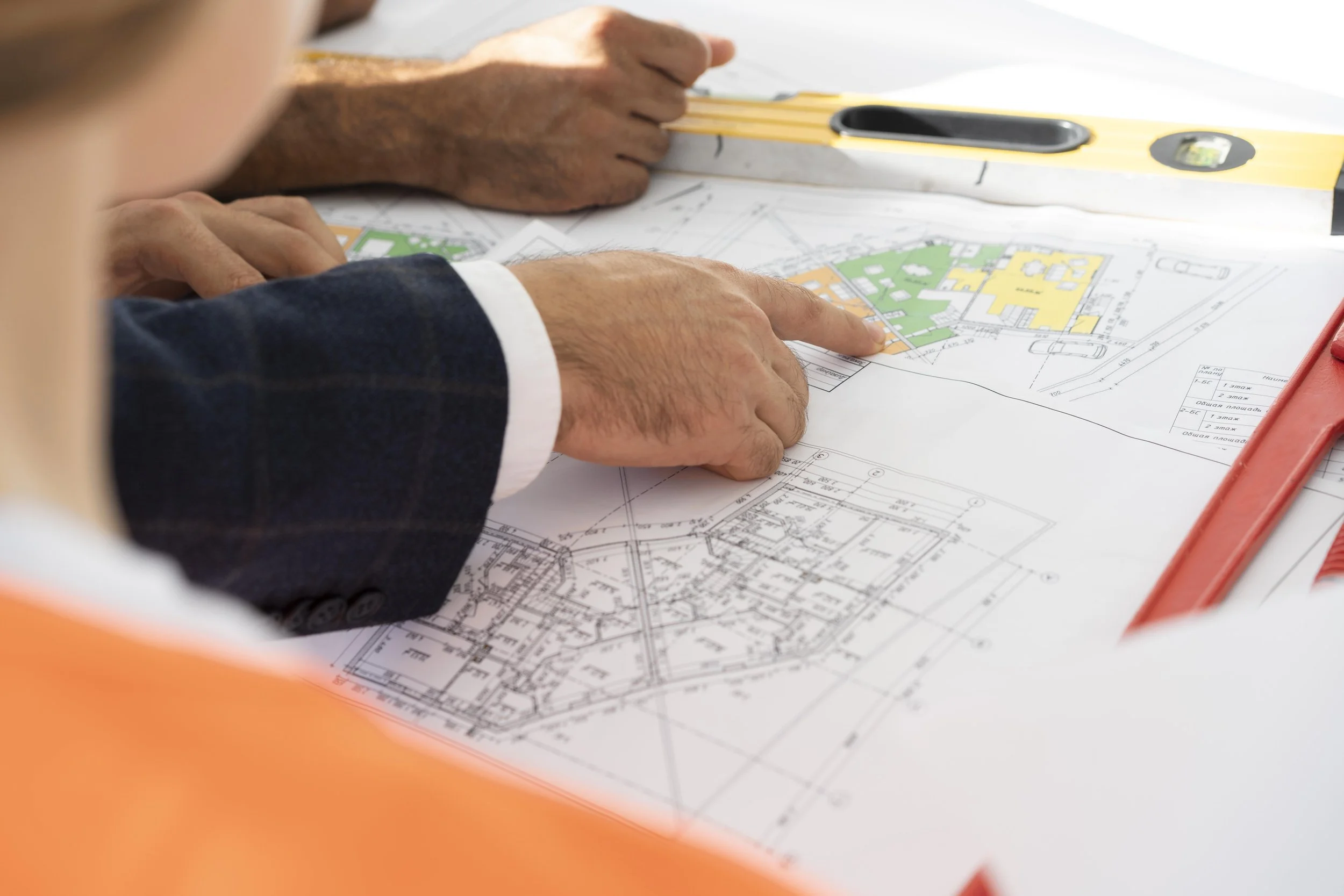 Two individuals reviewing architectural blueprints on a table, with one pointing at a specific area. Construction tools and a level are also present on the table.
