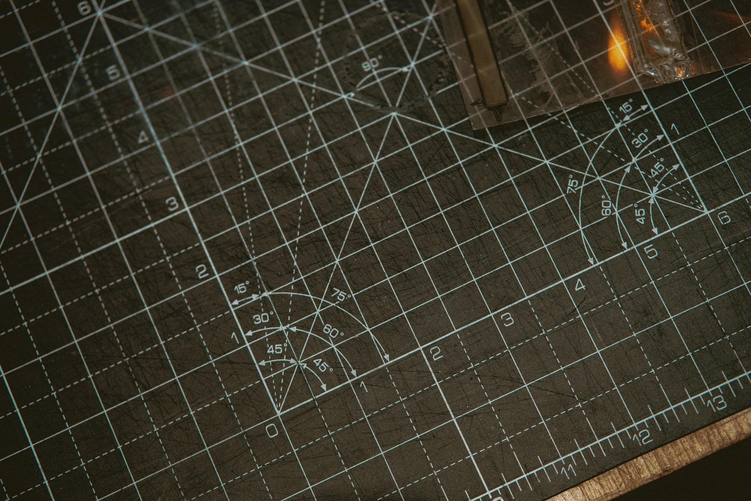 Close-up of a black cutting mat with white grid lines, measurements, and angle markings, with a small glass container holding a clear liquid and a metal tool resting inside.