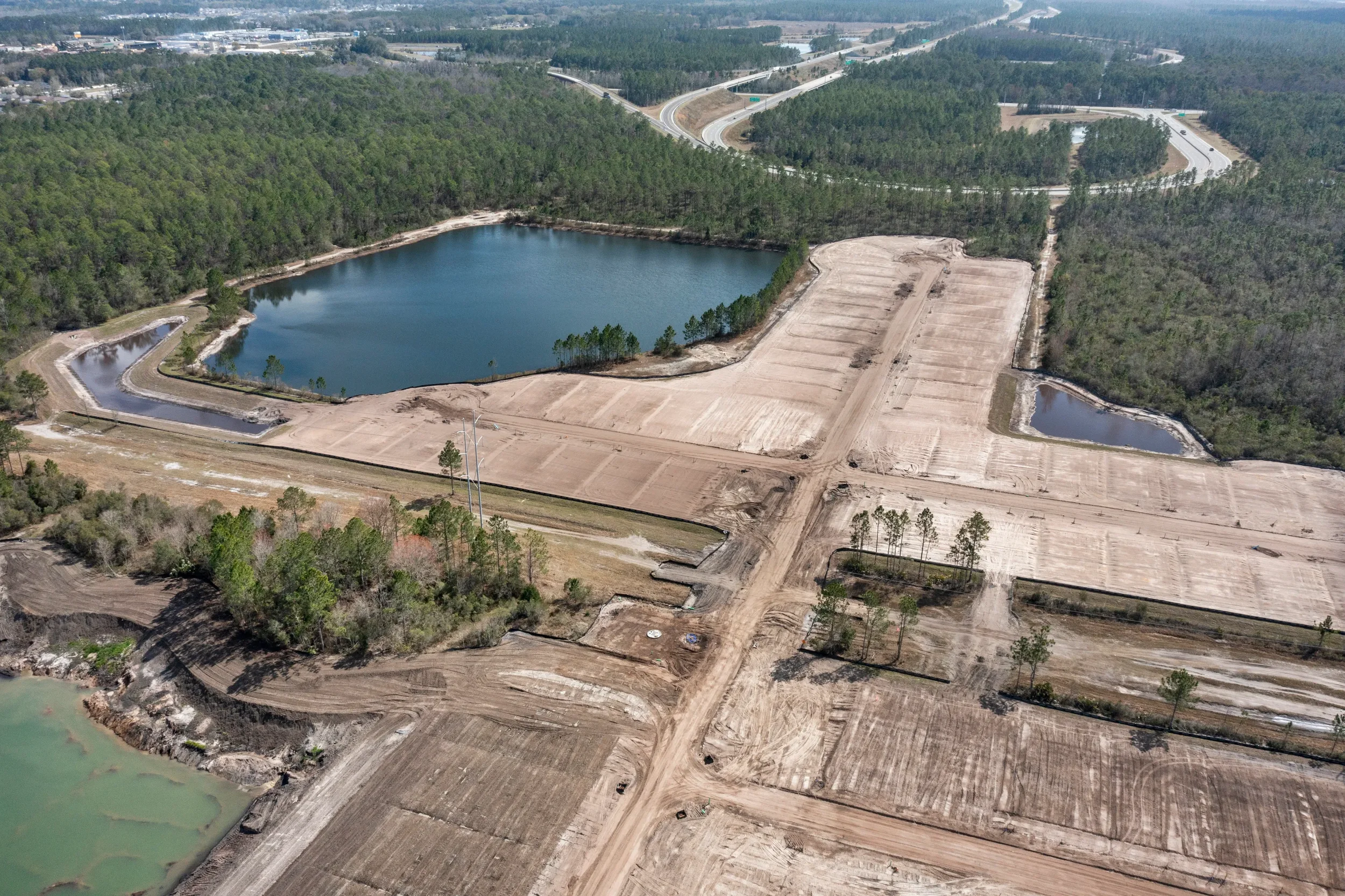 Aerial view of a construction site with cleared land, new roads, and small ponds, surrounded by dense green trees and forest, with a highway visible in the background.