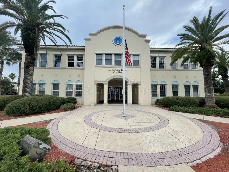 Jacksonville Beach City Hall, with an American flag on a flagpole in front, surrounded by palm trees and landscaped bushes.