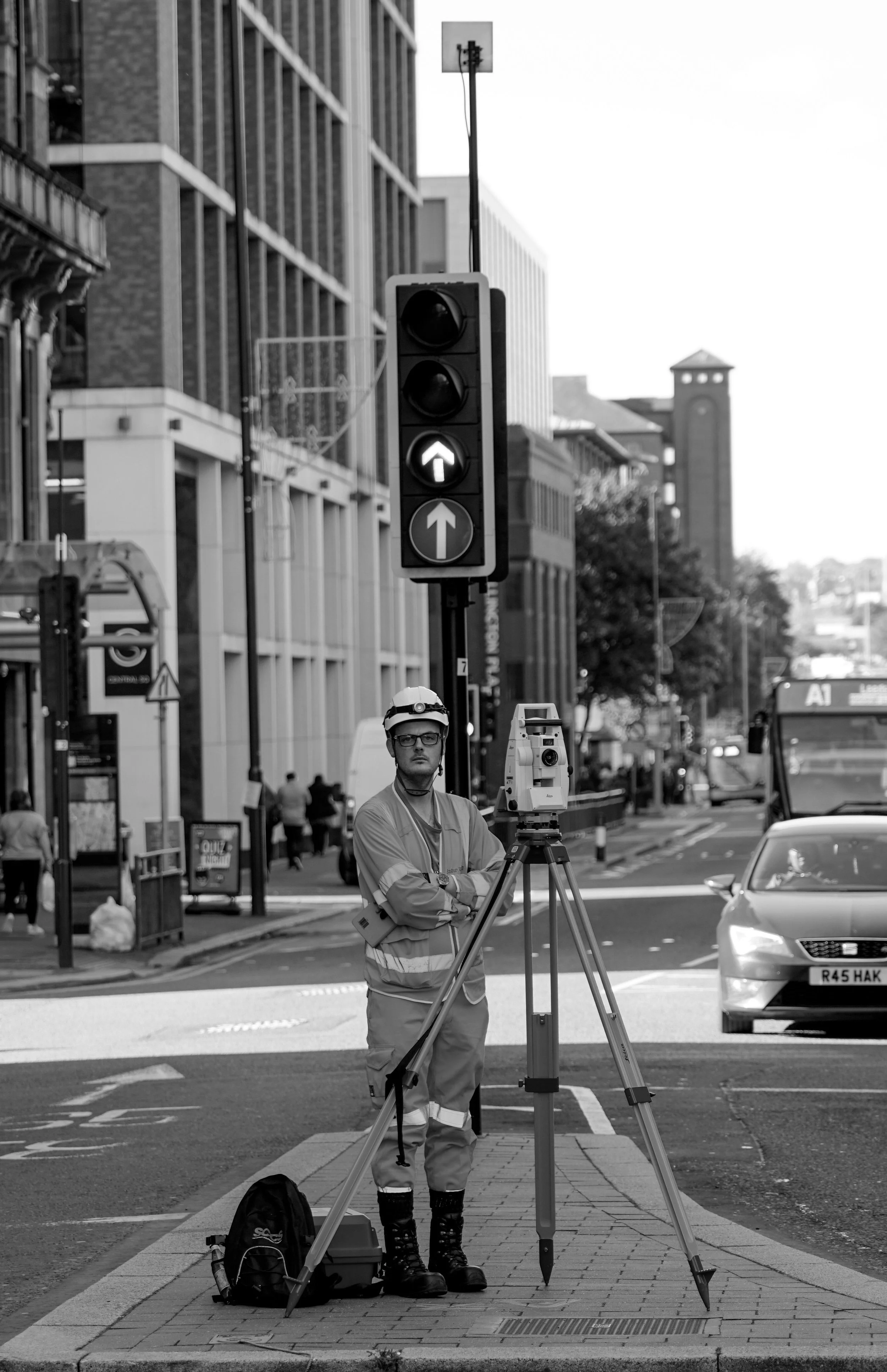 A surveyor wearing safety gear stands next to surveying equipment on a city street with modern buildings, vehicles, and pedestrians in the background.