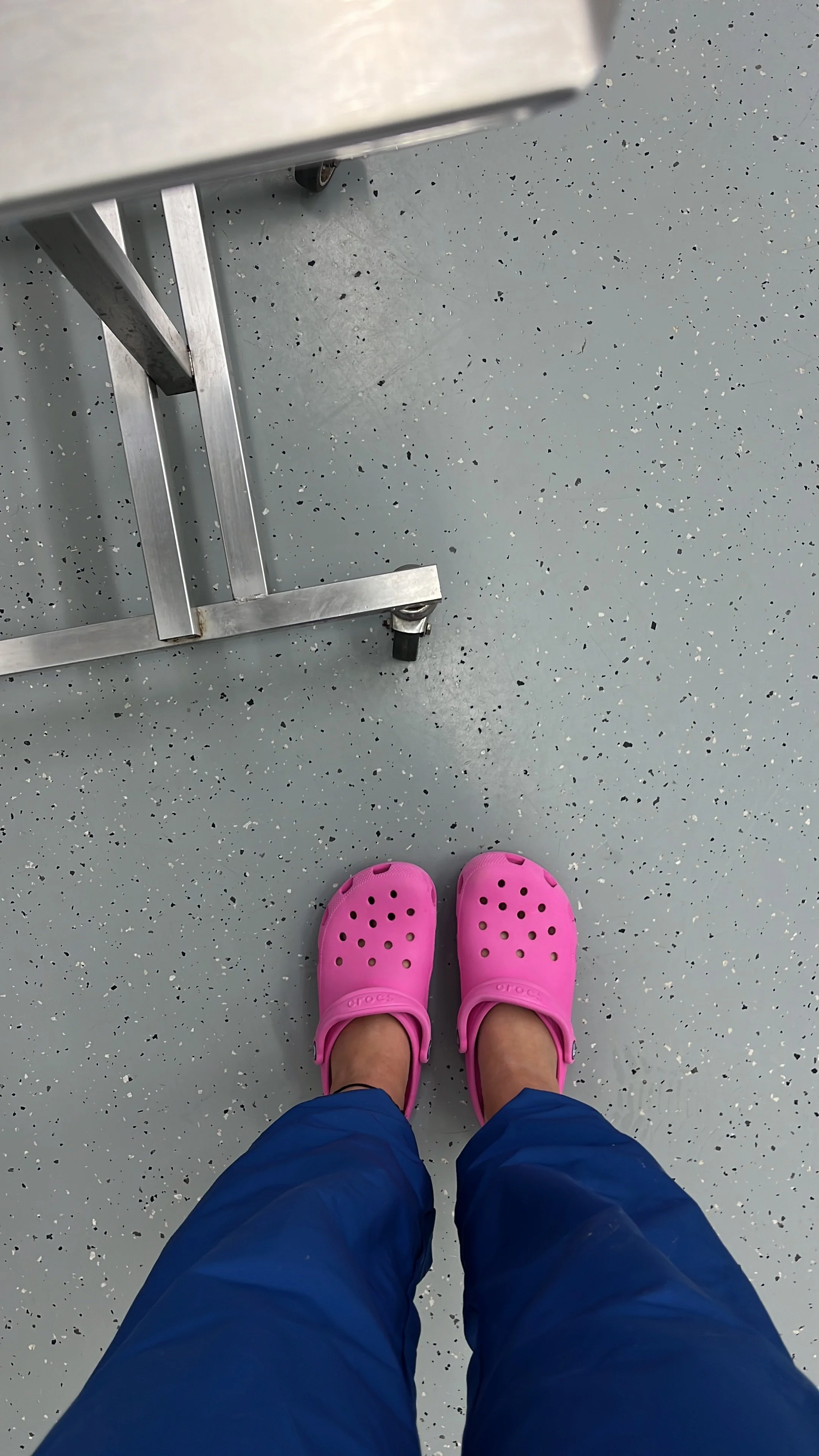 Blick nach unten auf eine Person in blauen Hosen und pinken Crocs auf einer grauen, gepunkteten Bodenfliese, daneben ein Stehtisch auf Rollen.