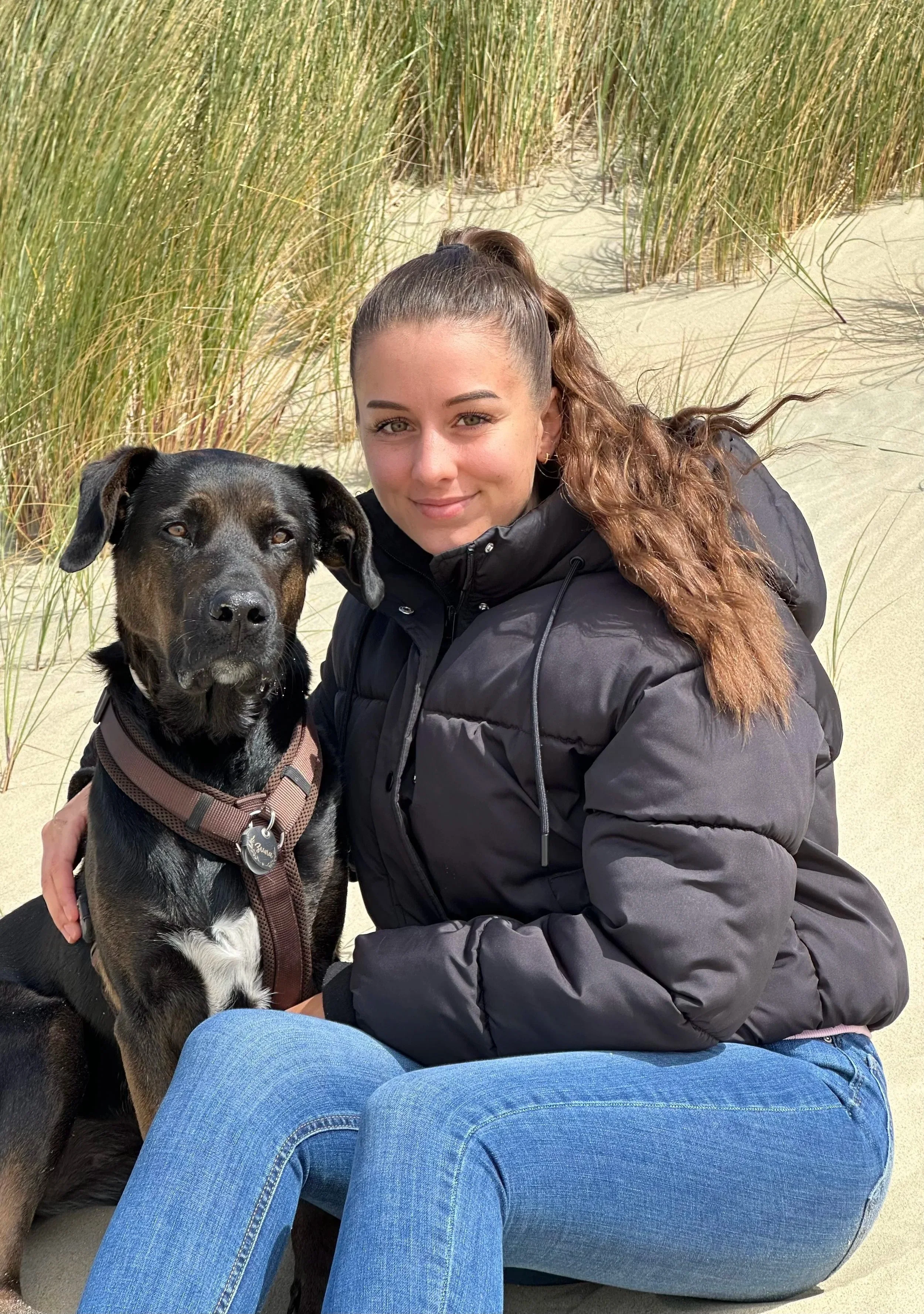 Junge Frau mit braunen, lockigen Haaren in einem schwarzen Jacke sitzt im Sand an einem Strand, umgeben von hohen Dünen und Gräsern, mit einem schwarzen Hund.