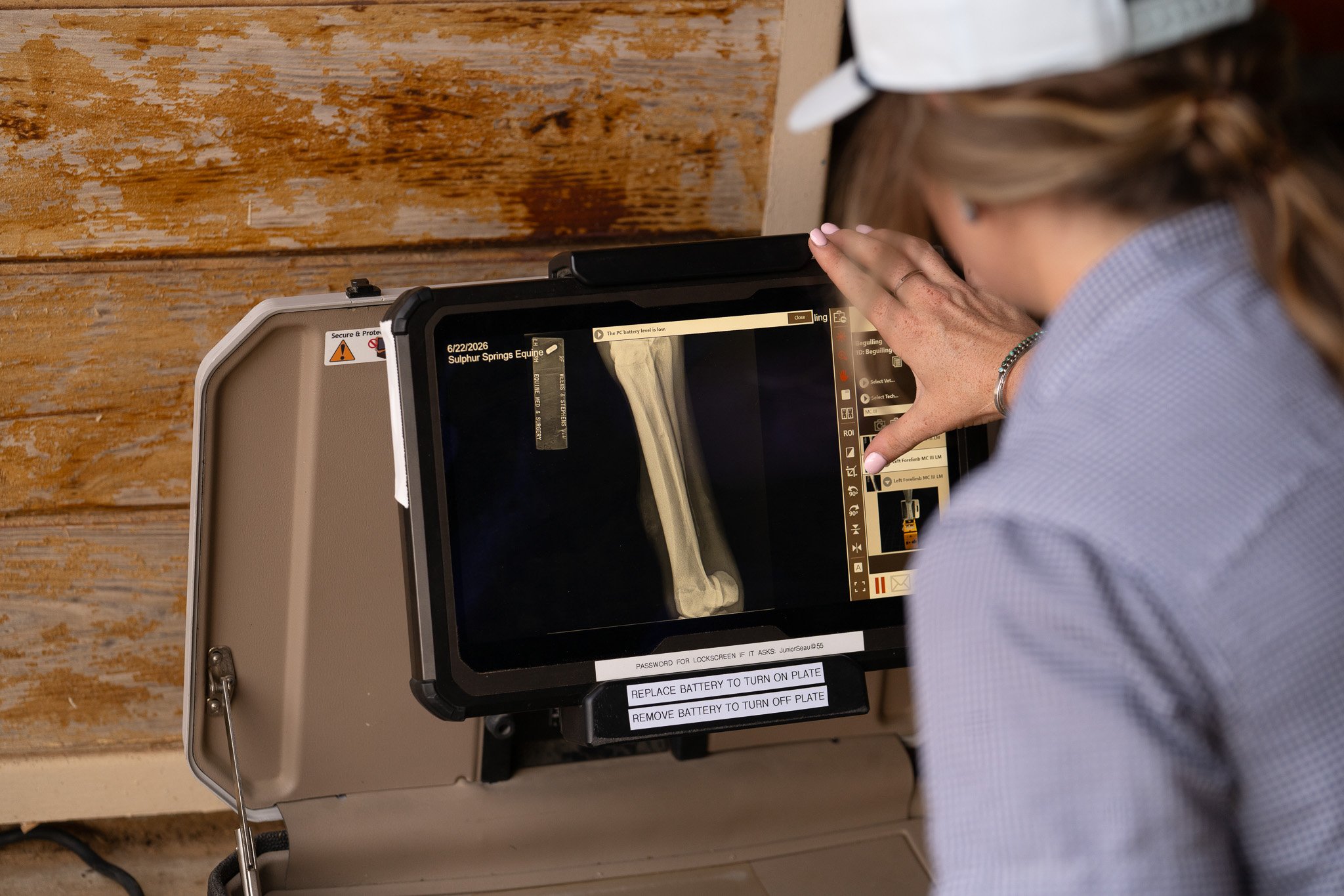 A person wearing a white cap and a checkered shirt is pressing a button on a portable ultrasound device, which displays an image of a horse's leg. The device is open and placed on a wooden surface.