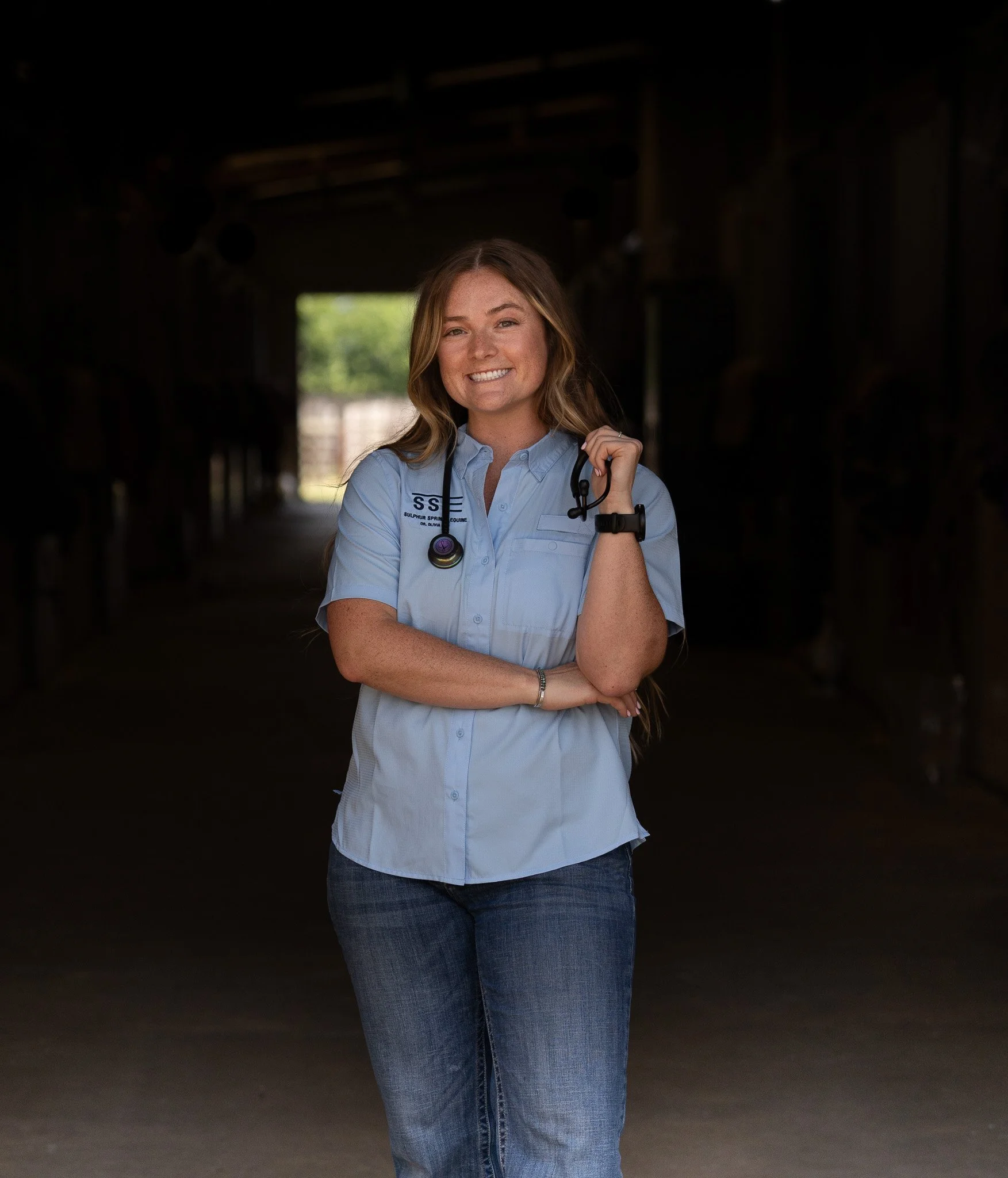A young woman with long, light brown hair smiling, dressed in a light blue shirt with a stethoscope around her neck and
