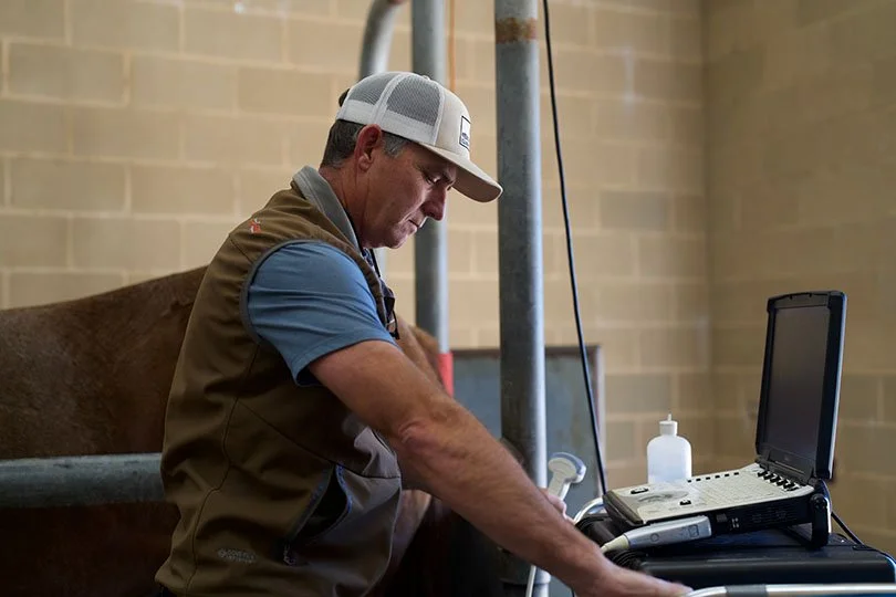 A man wearing a baseball cap and a brown vest using a laptop at a workstation with a bottle of hand sanitizer nearby.