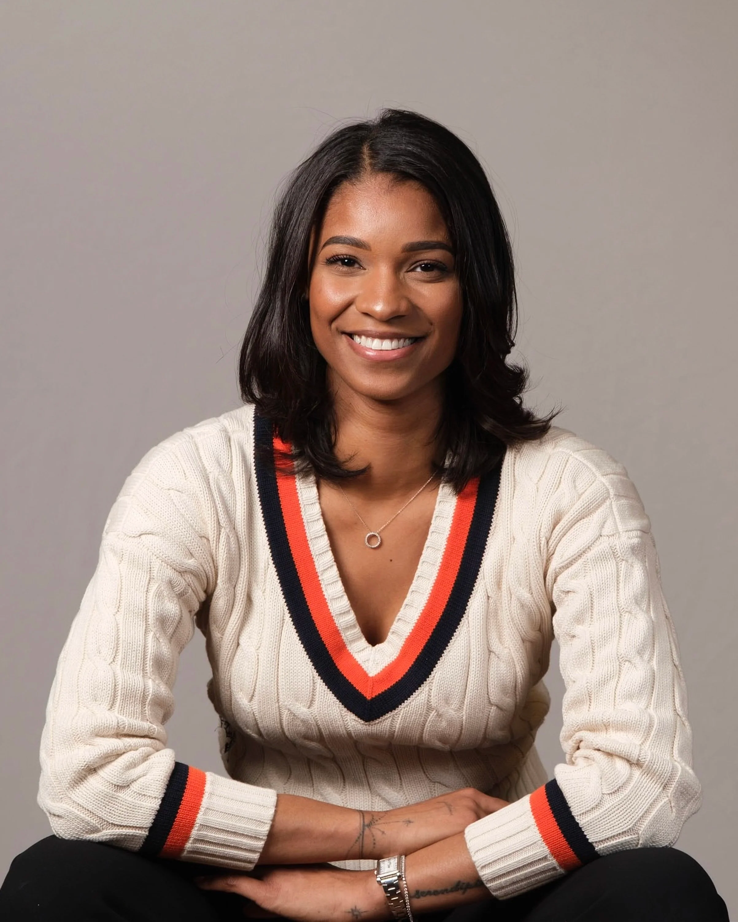 A woman with dark hair, smiling, wearing a cream-colored sweater with orange and black striped V-neck, black pants, a silver watch, and a delicate necklace, sitting with her arms crossed in front of a plain light grey background.