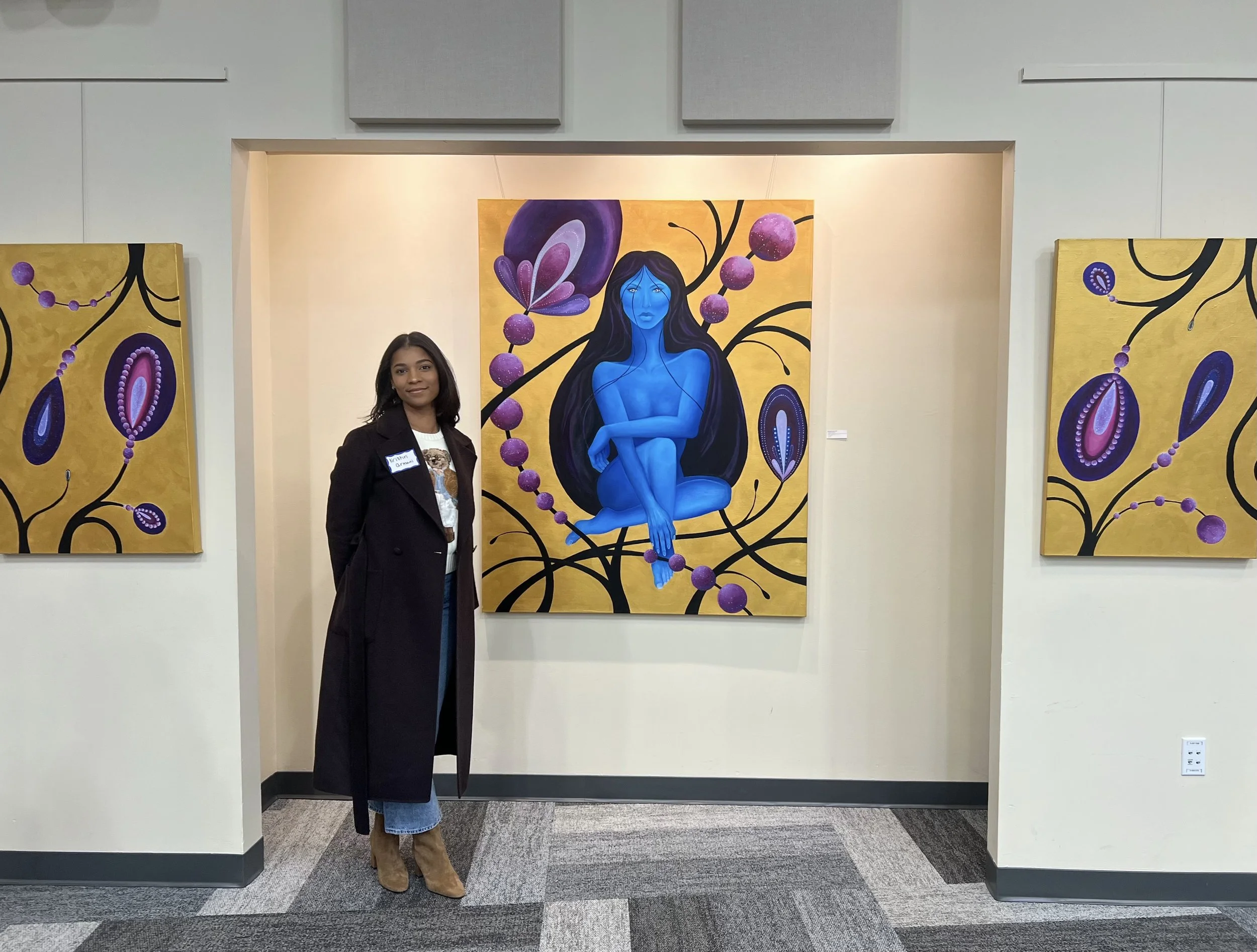 A woman standing in an art gallery in front of a colorful painting of a blue-skinned woman with long black hair, surrounded by purple and pink abstract floral designs.