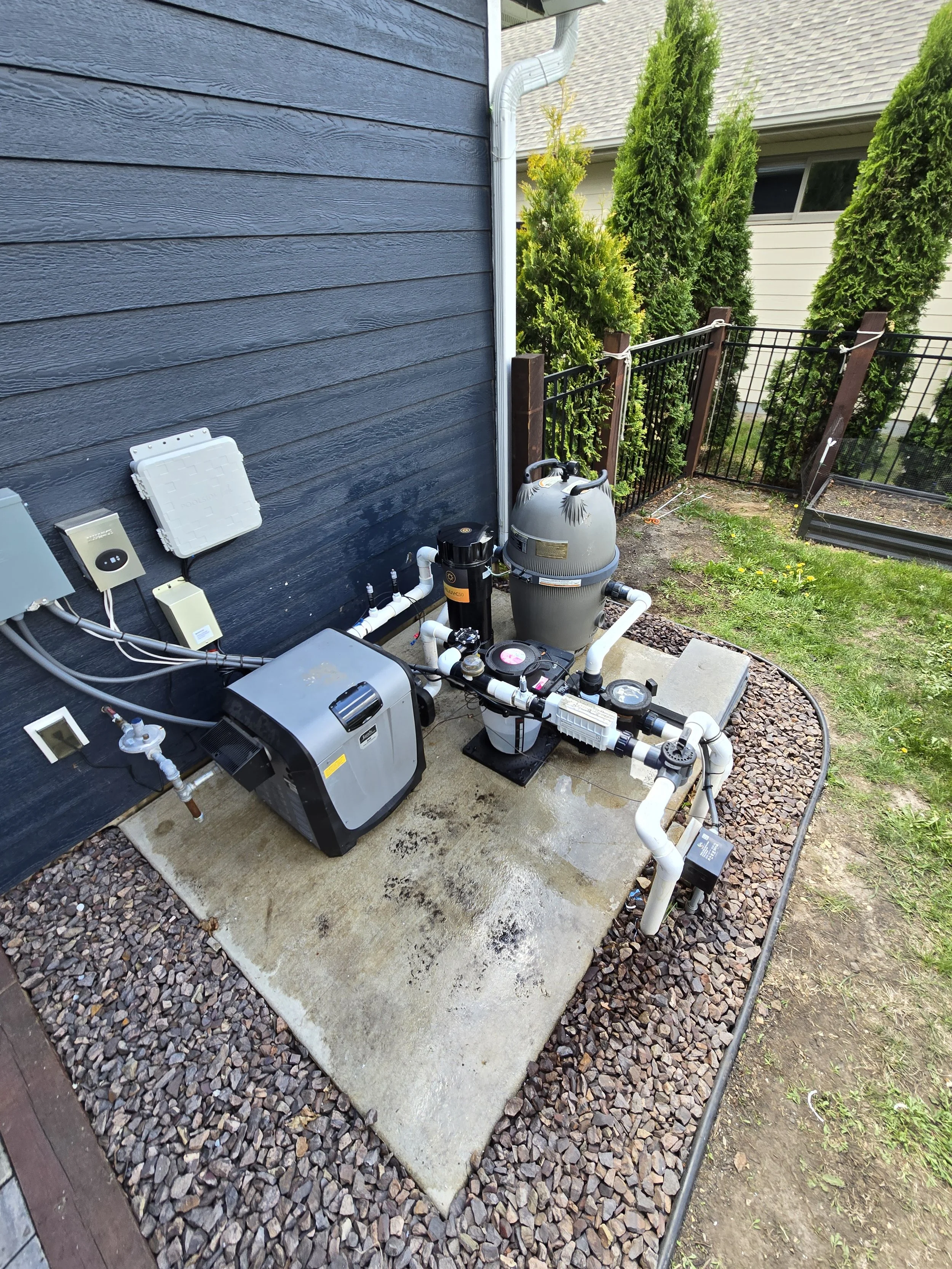 Installation of a pool filtration system next to a black house wall, with green trees and a yard in the background.