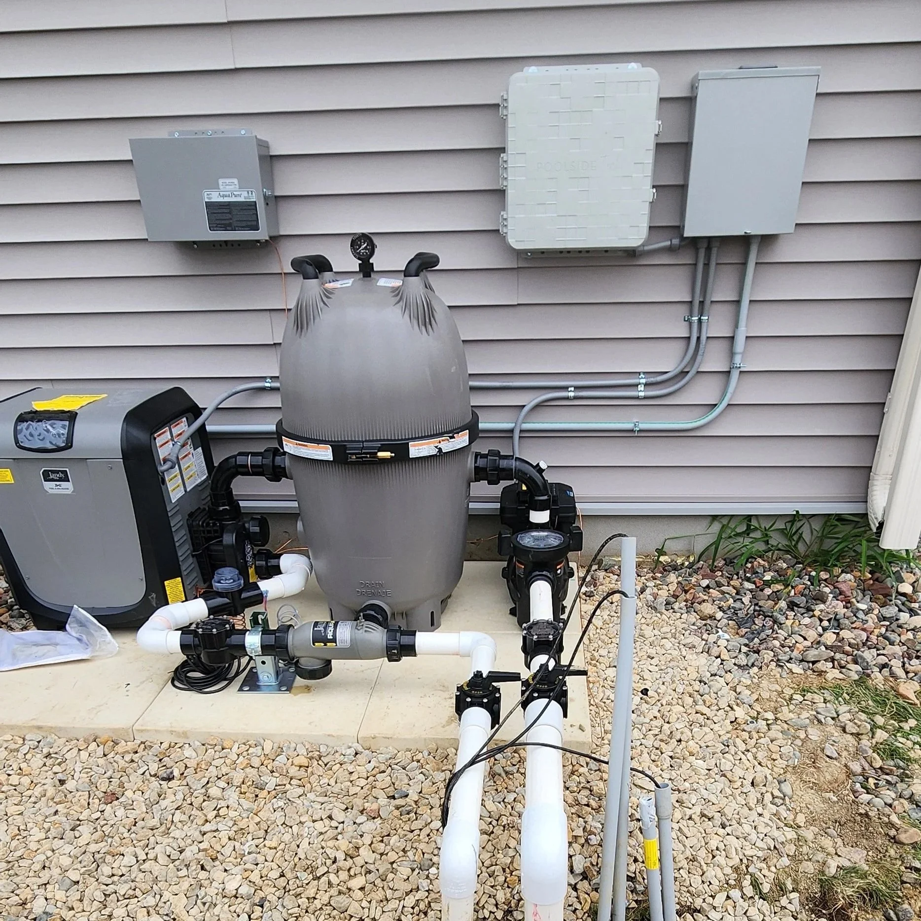 Outdoor pool equipment setup with a large gray filter tank, pressure gauges, interconnected pipes, and electrical boxes mounted on beige house siding, with gravel and small plants on the ground.