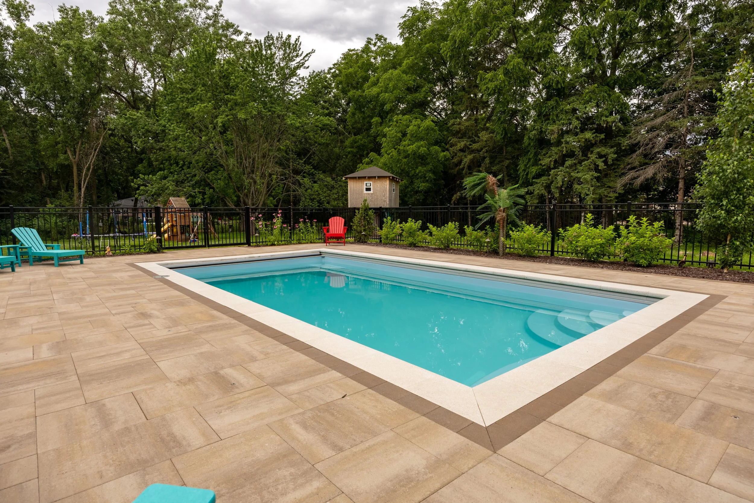 A backyard with a swimming pool, surrounded by a beige tile deck and a black fence. There are four colorful Adirondack chairs and a small garden with blooming flowers and various plants. In the background, tall green trees and a wooden playhouse or shed are visible, with cloudy skies overhead.