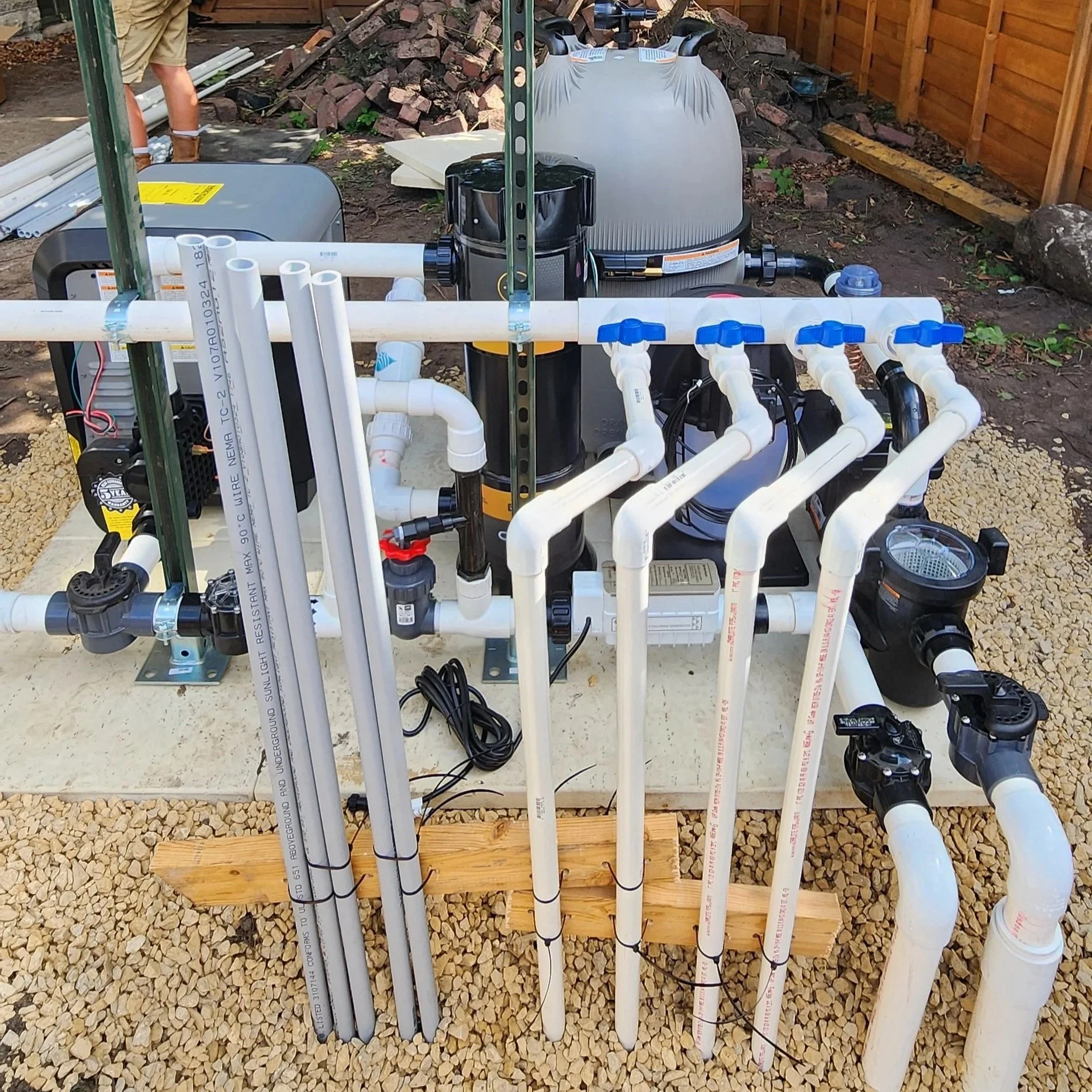 An outdoor water filtration system on a gravel surface with white PVC pipes and valves, a black pump, a large gray water tank, and construction materials and a wooden fence in the background.