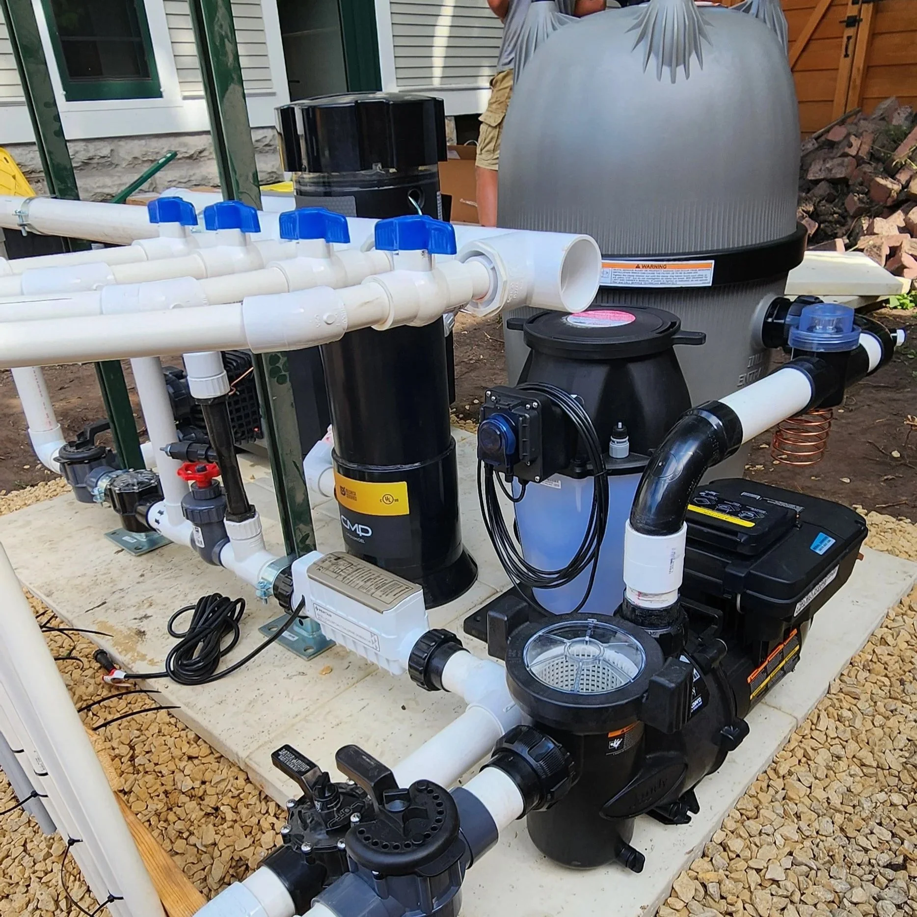 An outdoor pool filtration system with white PVC pipes, black cylinders, and a large gray sand filter, installed on a concrete platform with gravel surrounding it, in a backyard with a house, fence, and tools in the background.