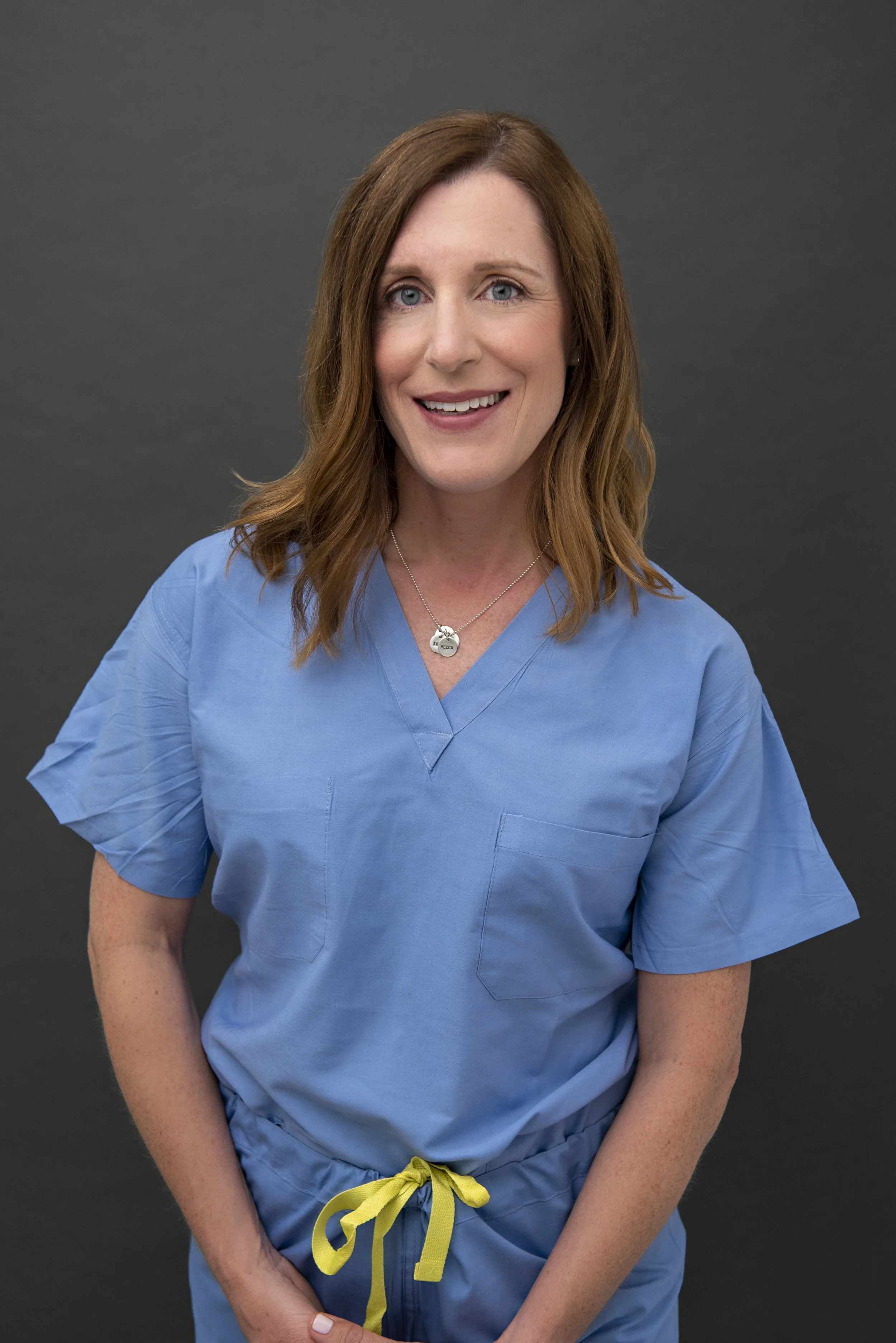 A woman with red hair and blue eyes wearing blue medical scrubs and a silver necklace, standing against a dark gray background, smiling at the camera.