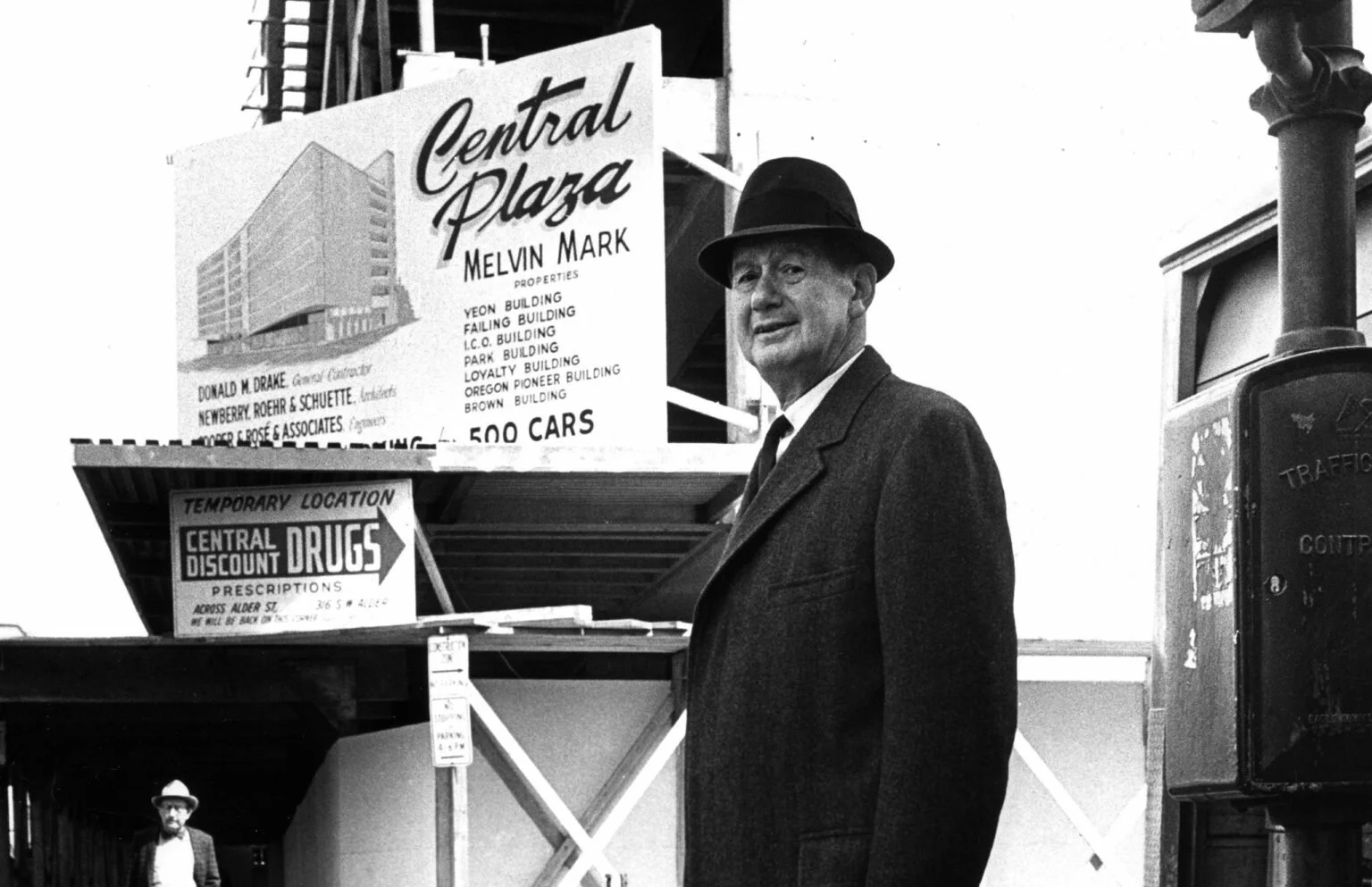 A man wearing a dark suit and hat stands in front of a 'Central Plaza' sign with various building names. A smaller sign for 'Central Discount Drugs' is below. Another man wearing a hat and glasses is in the background, under an overpass.
