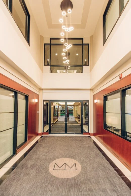 Modern hotel lobby with glass entrance doors, carpeted floor with logo, and high ceiling with hanging circular light fixtures.