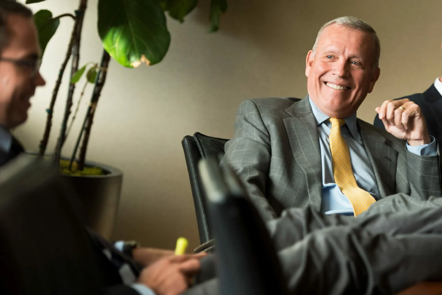 A group of businesspeople in a meeting room, with one man prominently smiling and wearing a gray suit and yellow tie.