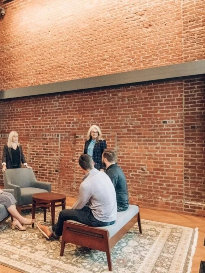 A group of four people in a cozy sitting area with exposed brick walls, a patterned rug, and modern furniture. Two women are standing, and two men are seated on a bench, engaged in conversation.