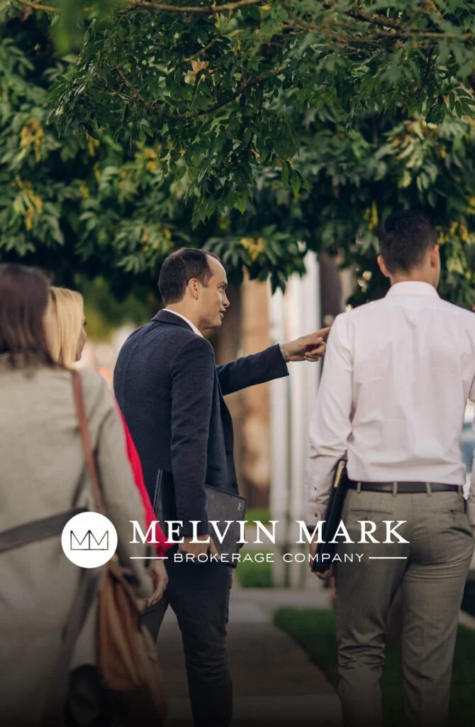 Businesspeople walking outdoors, with one man gesturing and holding a laptop, under green trees, promoting Melvin Mark Brokerage Company.