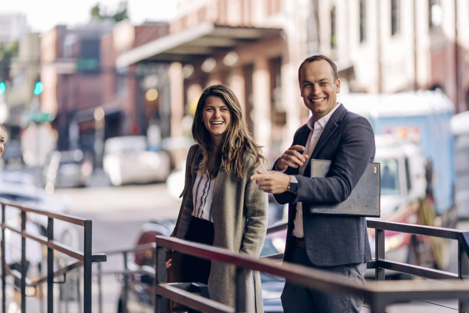 Two business professionals, a woman with long brown hair and a man with short brown hair, are smiling and talking outdoors on a city street, dressed in business attire.