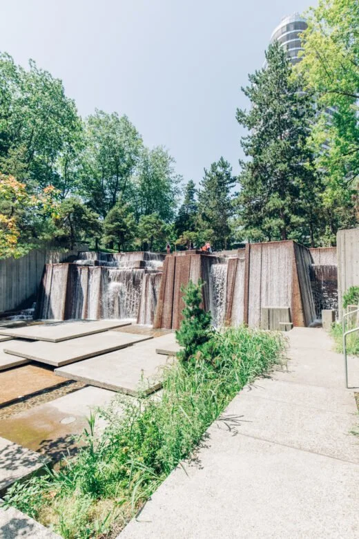 A small waterfall with cascading water over a series of rust-colored metal barriers, surrounded by green trees and plants, with a concrete walkway nearby.