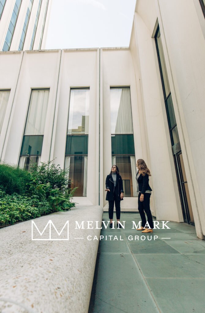Two women standing and talking outside a modern building with large windows and white exterior walls, next to a planter with green plants.