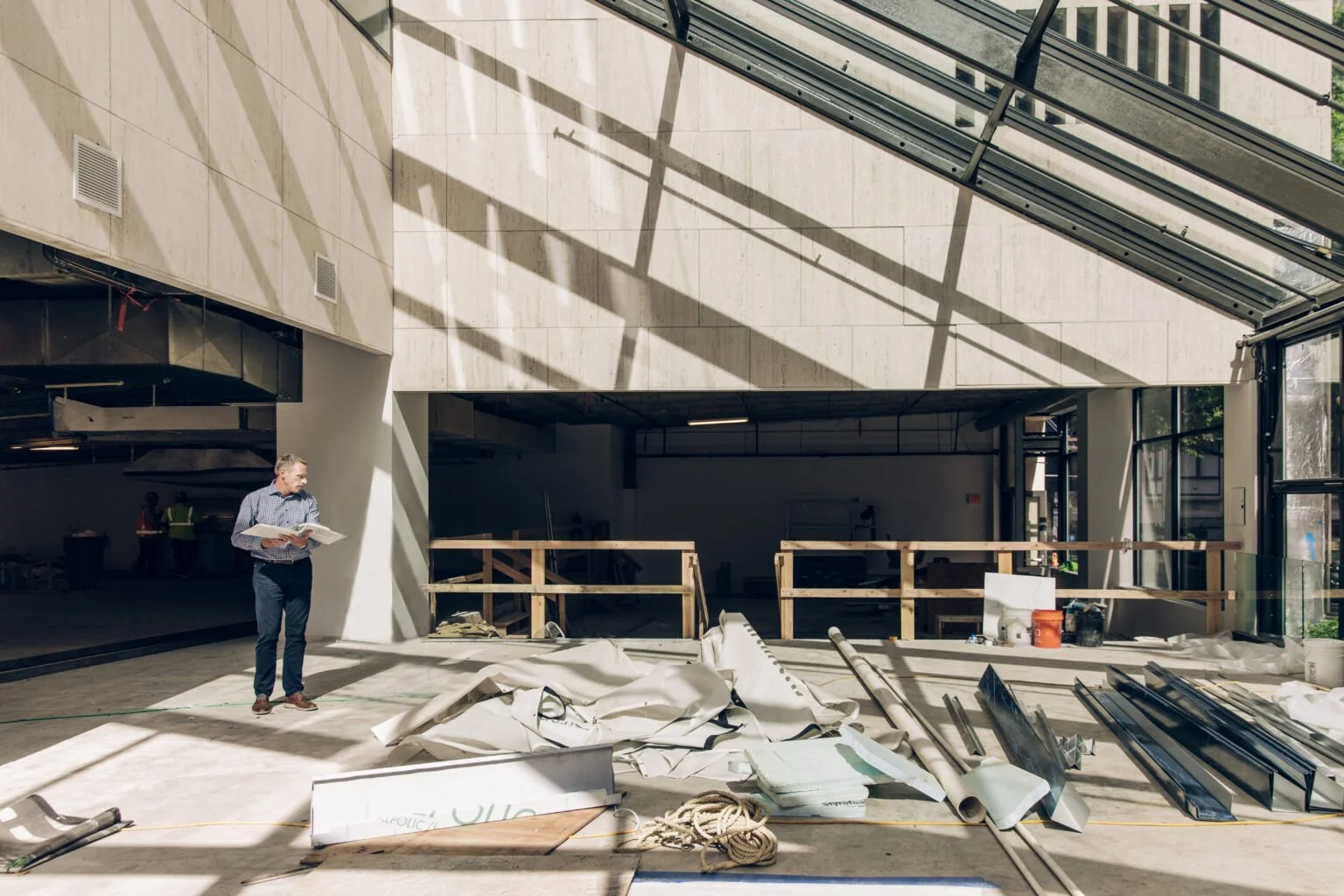Interior of a building under construction, with a man standing on the left holding a notebook, construction materials and tools scattered on the floor, and sunlight casting shadows through large windows.