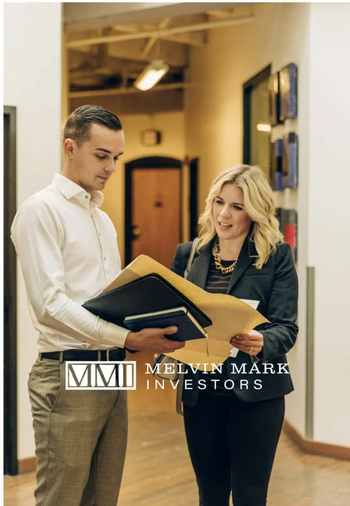 A young man and woman conversing in an office hallway, both holding documents, with the MELVIN MARK INVESTORS logo at the bottom.