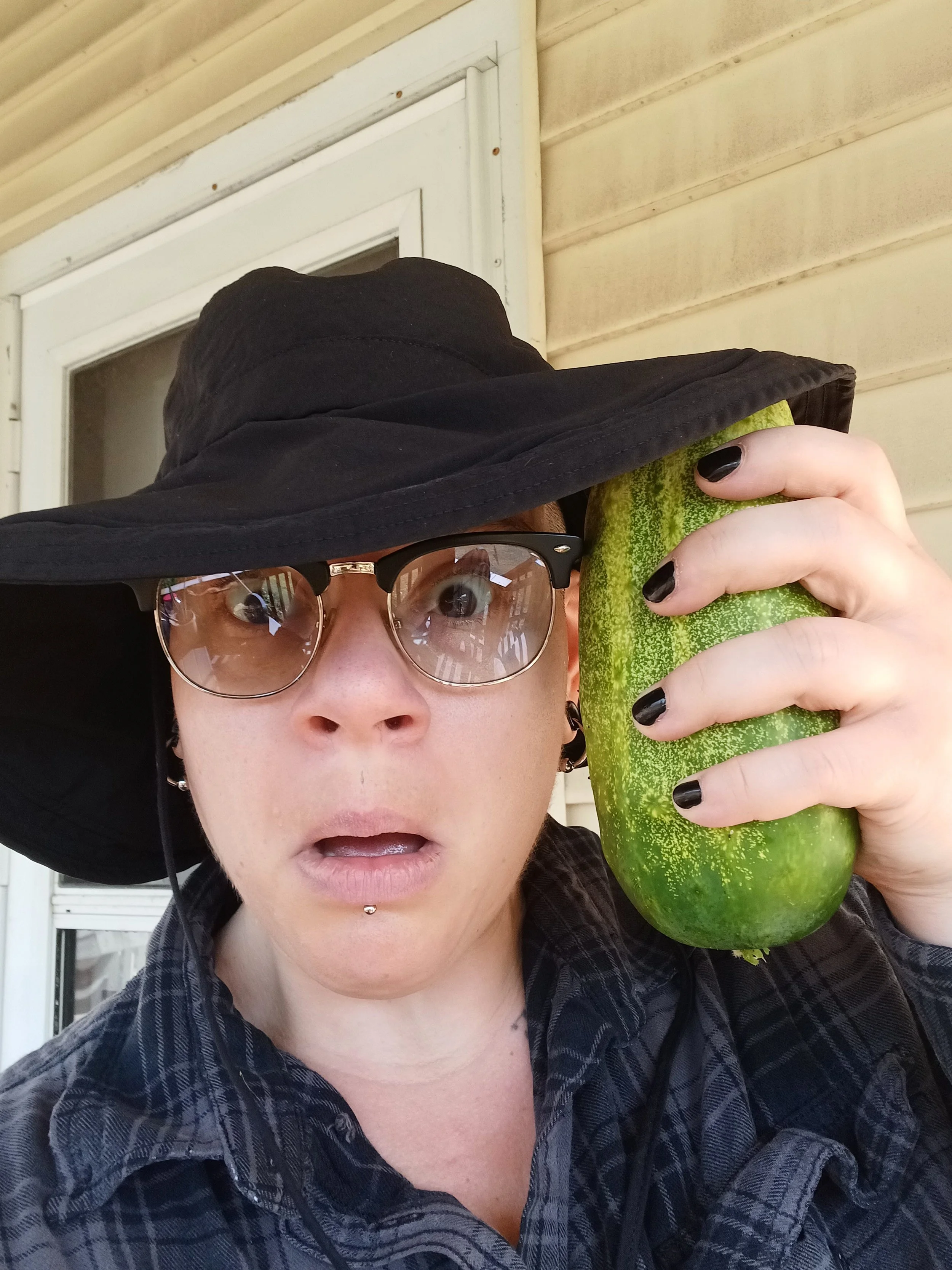 Person with glasses, a black wide-brim hat, and black painted nails holding a large cucumber close to their face.