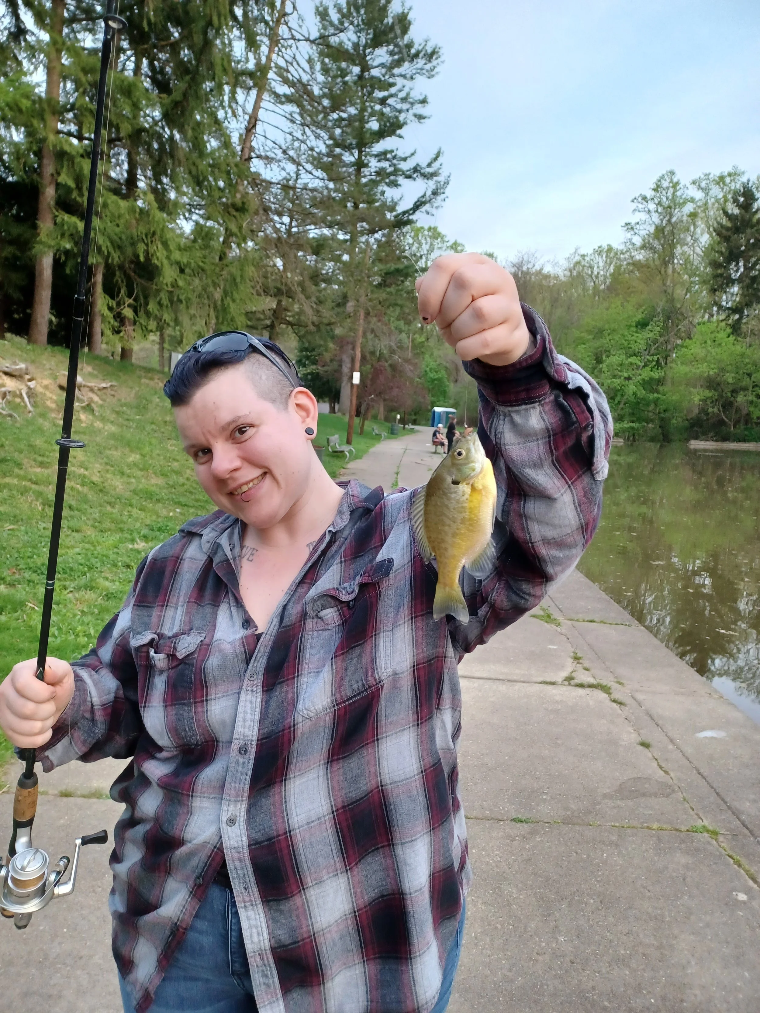 A smiling person with short dark hair, wearing sunglasses and a plaid shirt, holding a fishing rod and a fish they caught near a body of water in a park with trees and a paved path.