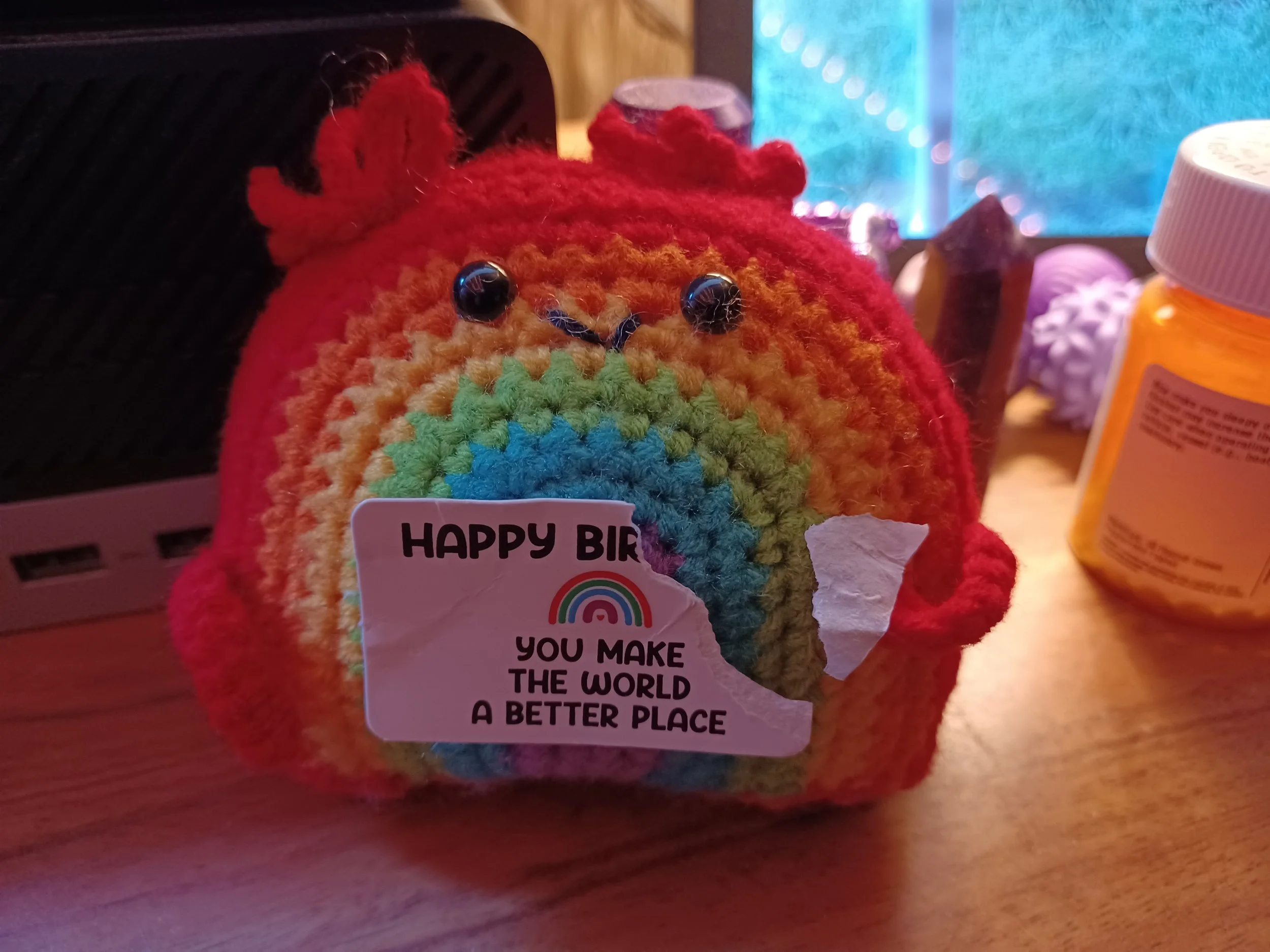 Colorful crocheted stuffed animal resembling a bird, with a rainbow sticker that says 'HAPPY BIRTHDAY' and a message underneath reading 'YOU MAKE THE WORLD A BETTER PLACE,' placed on a wooden surface surrounded by various containers.