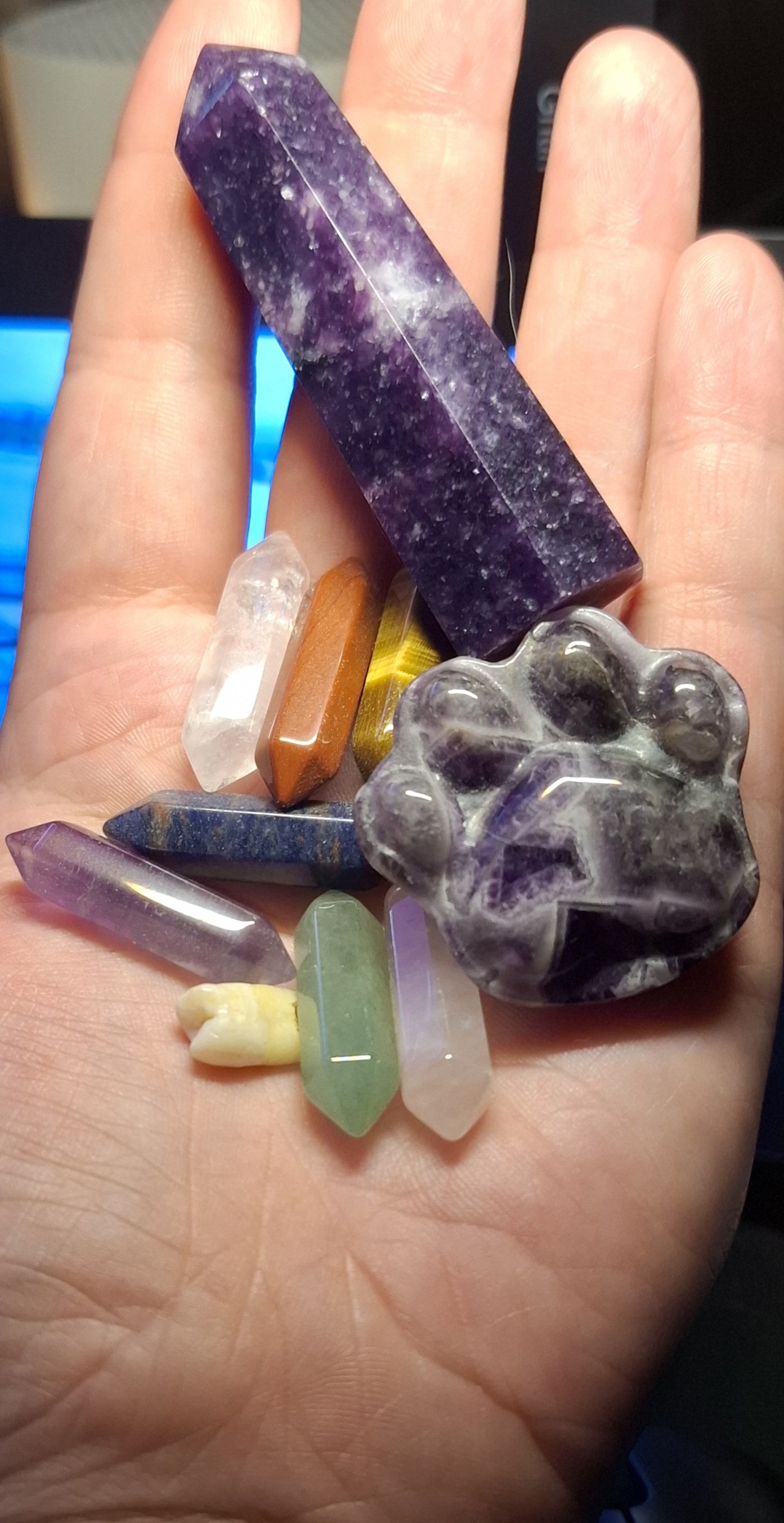 Hand holding a collection of colorful crystals and gemstones, including a large purple crystal, a clear quartz, and various other polished stones.
