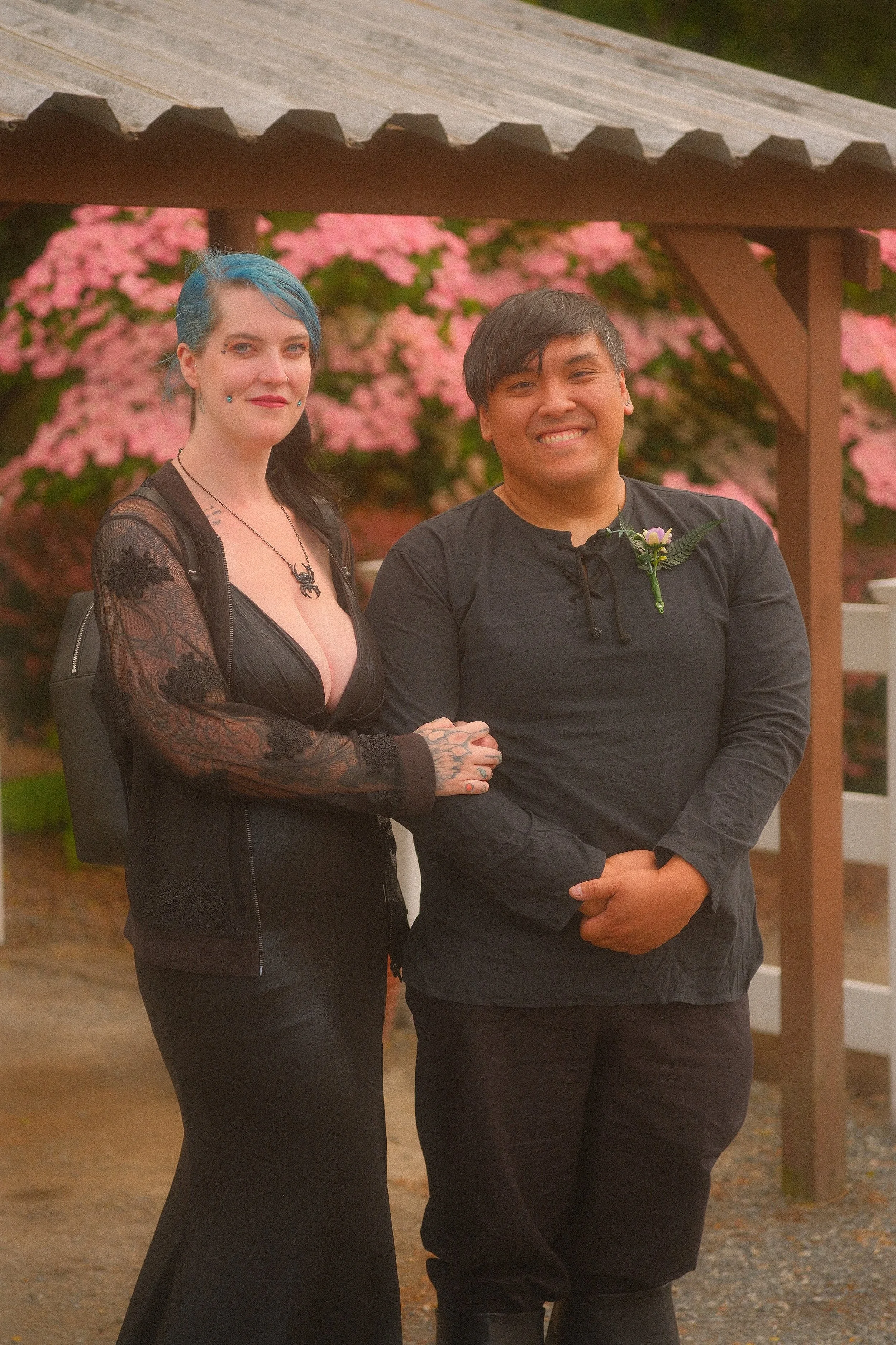 Two smiling people dressed in black pose together beneath a wooden gazebo surrounded by pink flowers.