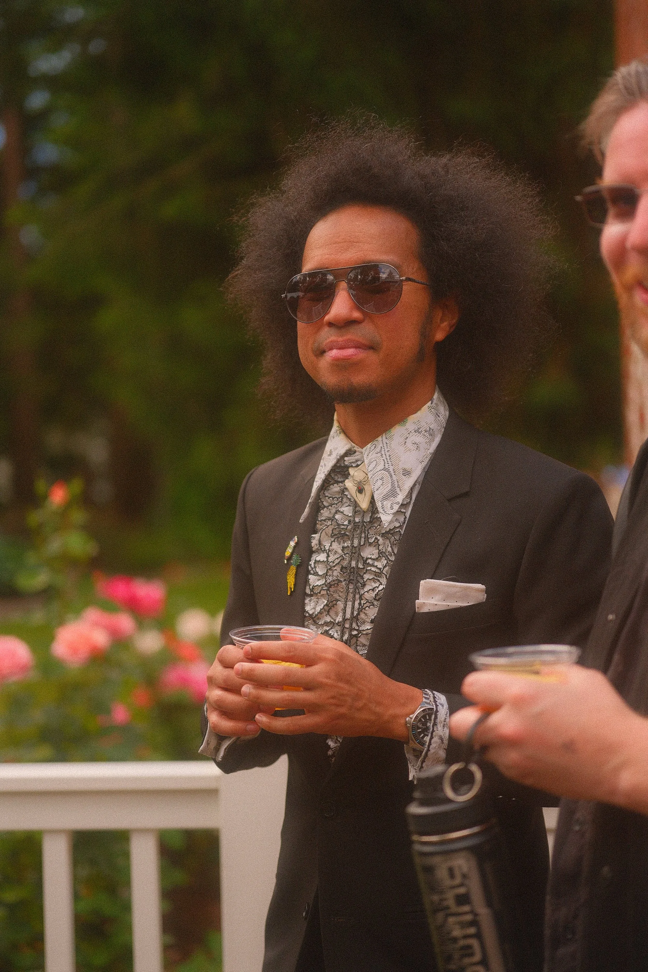 Person with an afro and aviator sunglasses wearing a black suit and patterned shirt, holding a drink at an outdoor event