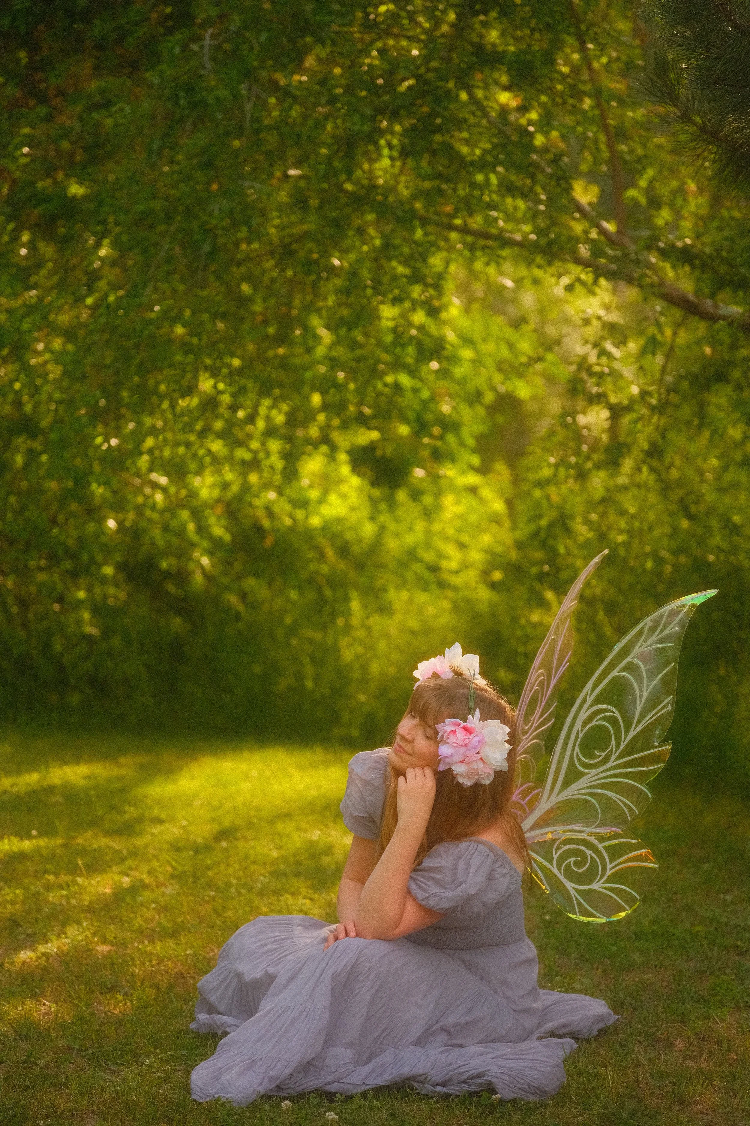 Young girl in a lavender dress wearing flower decorations and translucent fairy wings, sitting on grass in a sunlit garden.