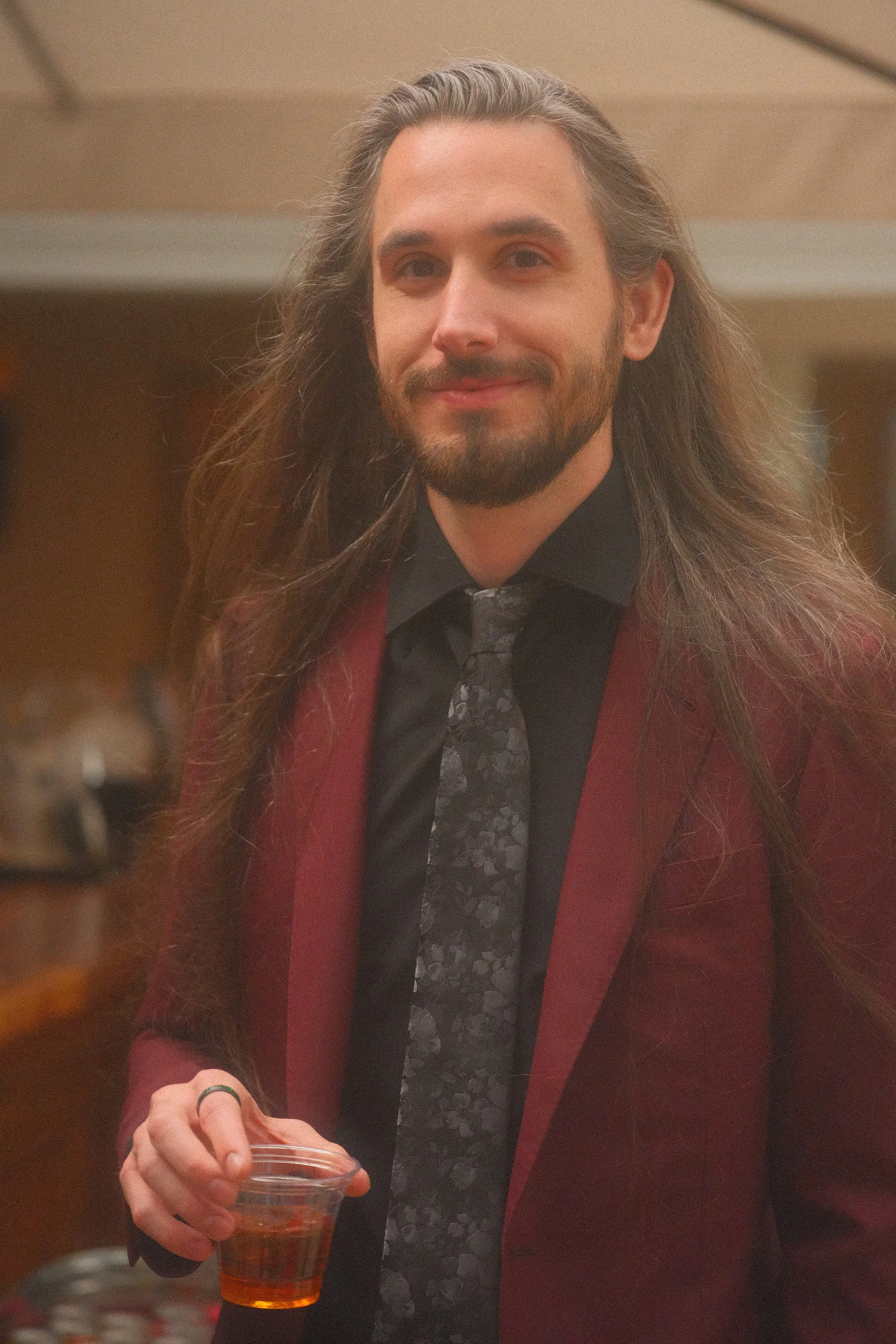 Smiling long-haired bearded man in a red coat and black scarf holding a glass of amber-colored drink