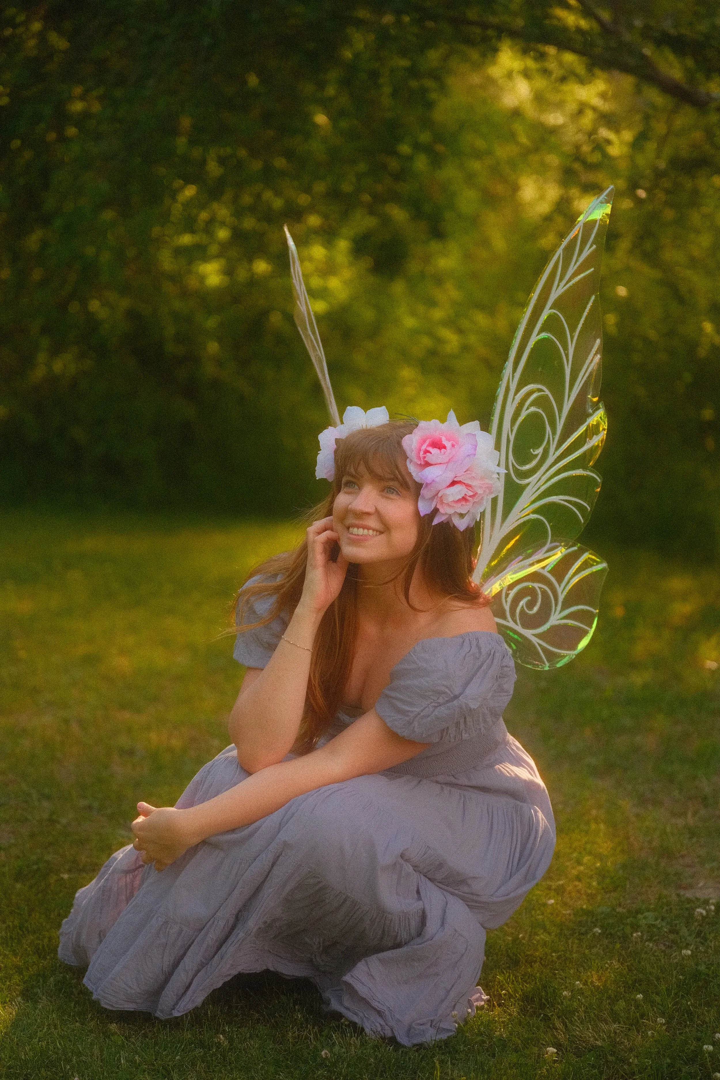 Smiling woman in a lavender dress and floral crown sitting on grass beside a decorative butterfly wing prop
