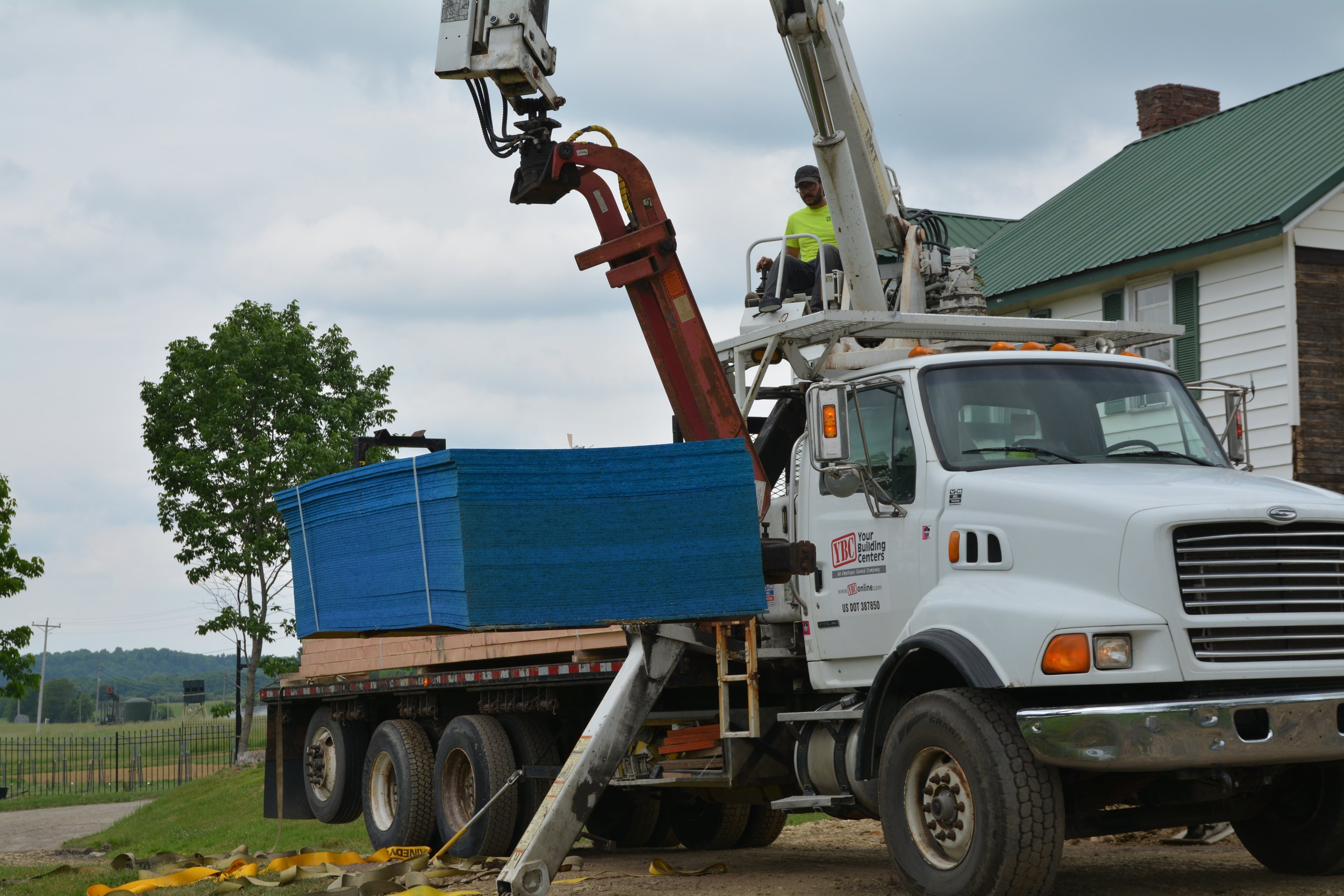 A worker operating a cherry picker lift on top of a truck, with the worker wearing a neon yellow shirt, in a residential area with a house having a green roof and trees in the background.