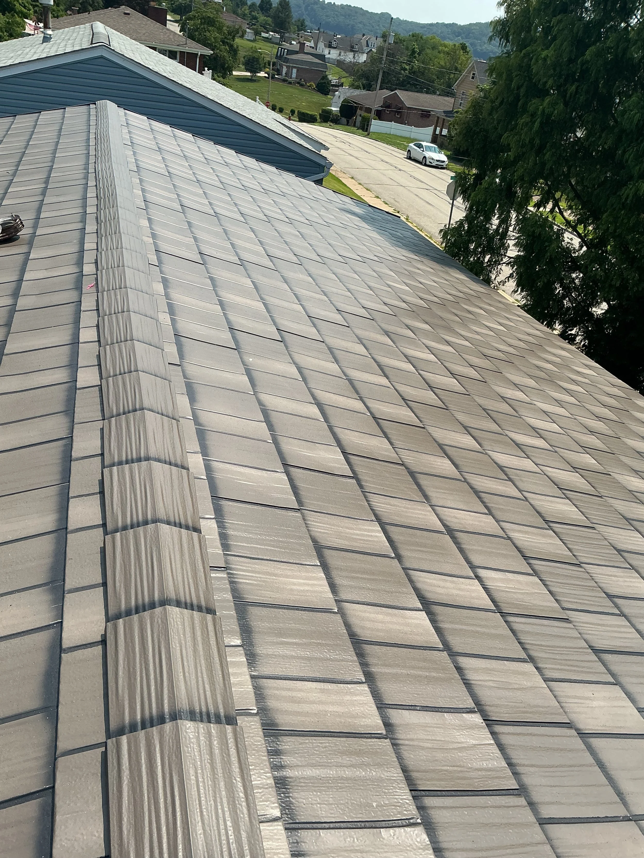 Roof with gray shingles and a ridge vent, overlooking a suburban neighborhood with houses, trees, and a street with parked cars.