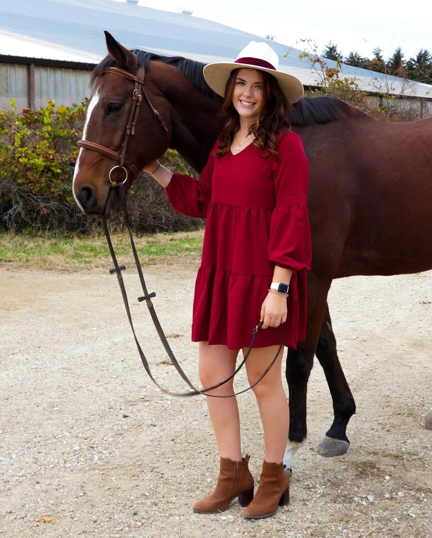 A woman in a red dress and brown boots standing next to a brown horse with a bridle, outdoors on a gravel surface. She is smiling and wearing a wide-brimmed hat.