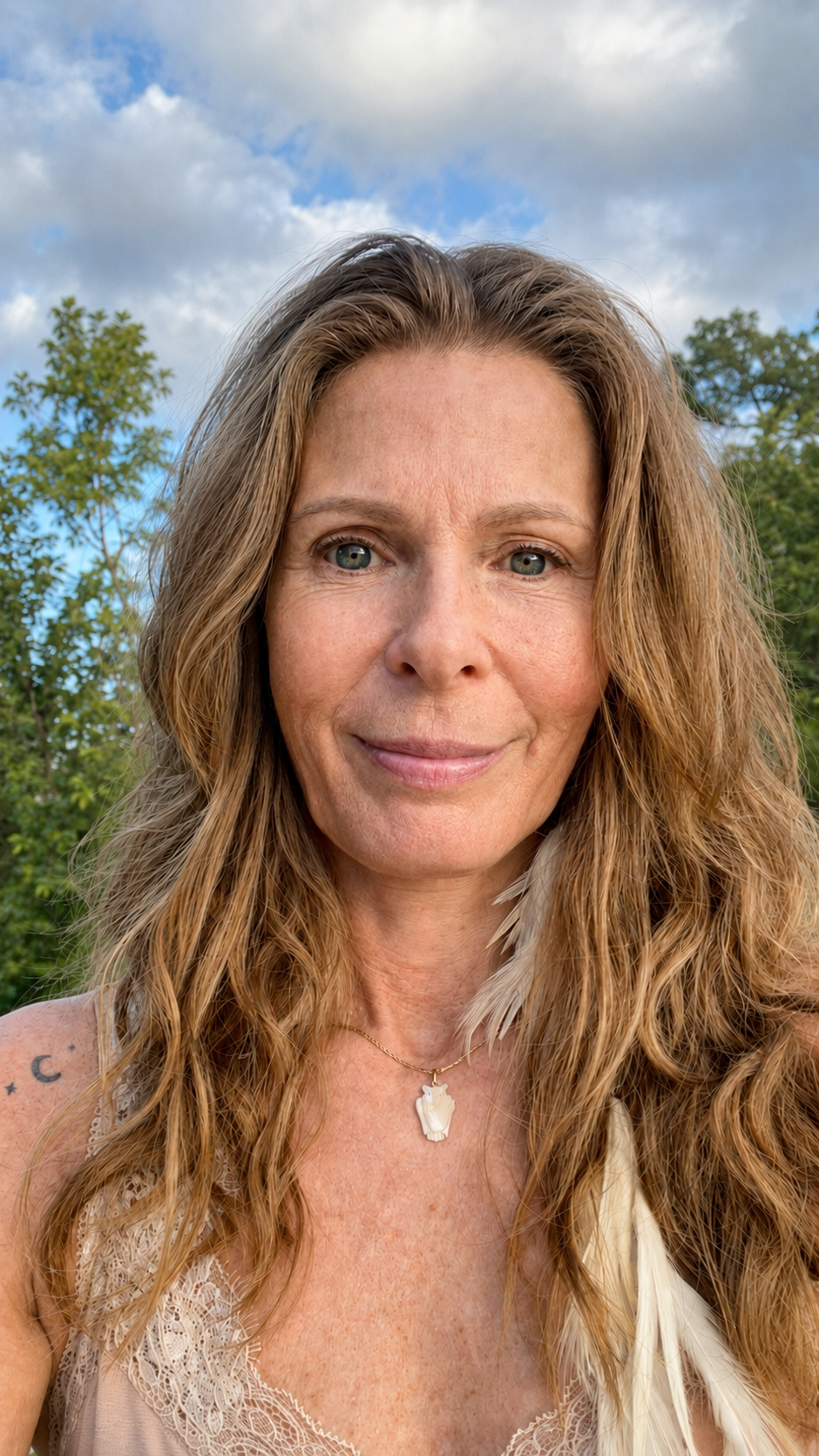 Een close-up van een vrouw met lange, golvende, lichtbruine haar, blauwe ogen en een lichte glimlach, gedragen in een heldere lucht met wolken en groene bomen op de achtergrond.