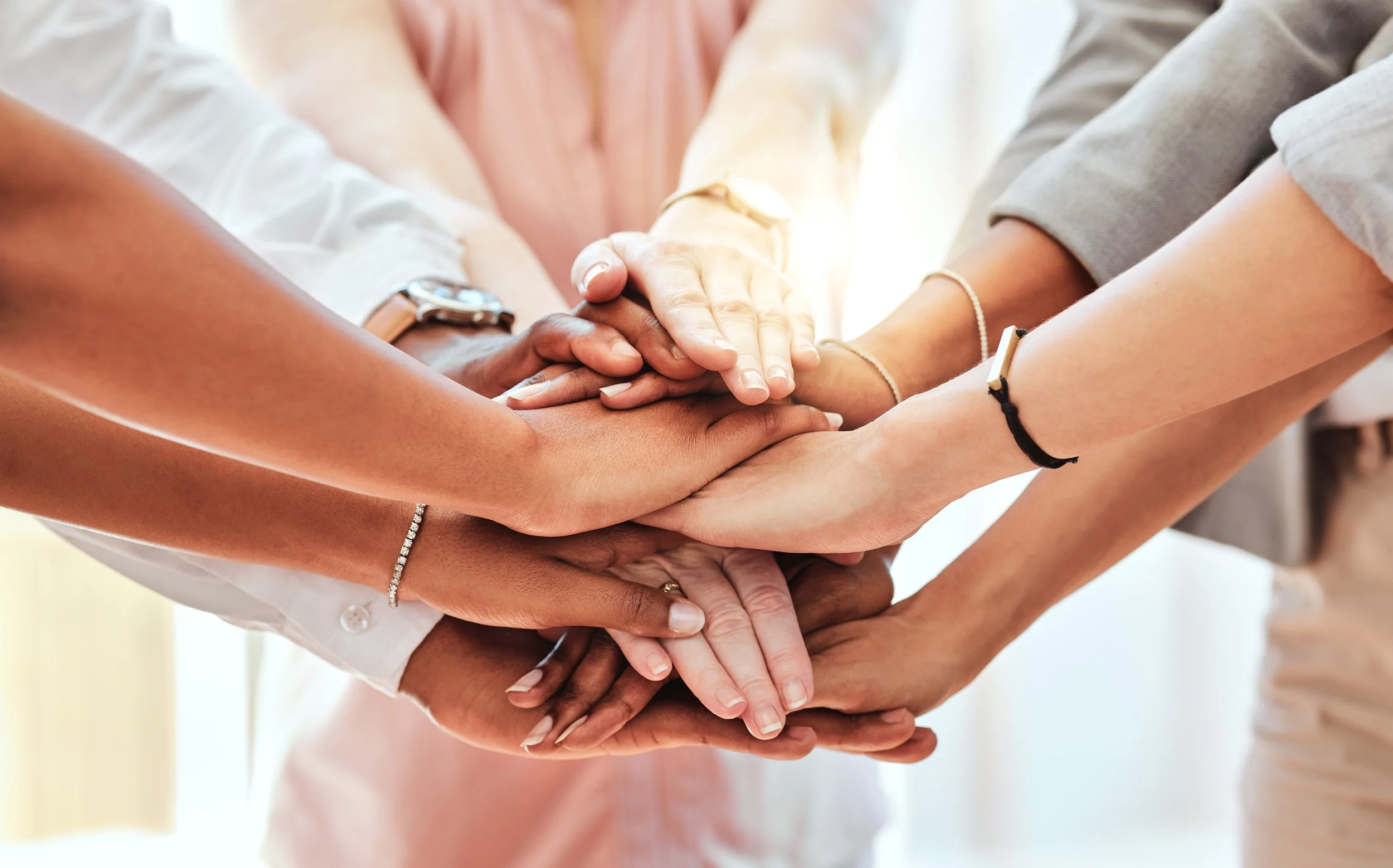 Multiple people placing their hands together in a group gesture of unity, showing diversity in skin tones and wearing bracelets and watches.