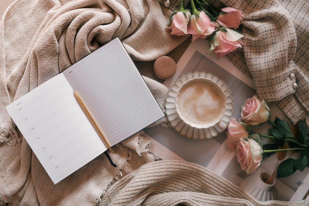Open planner with gold pen, pink roses, a cup of coffee, and soft blankets or fabrics.