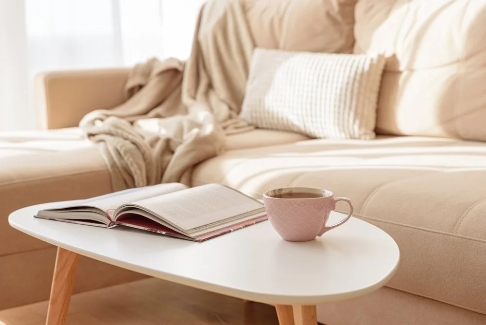 A cozy living room scene with an open book and a pink mug on a small white coffee table in front of a beige sofa with pillows and blankets.