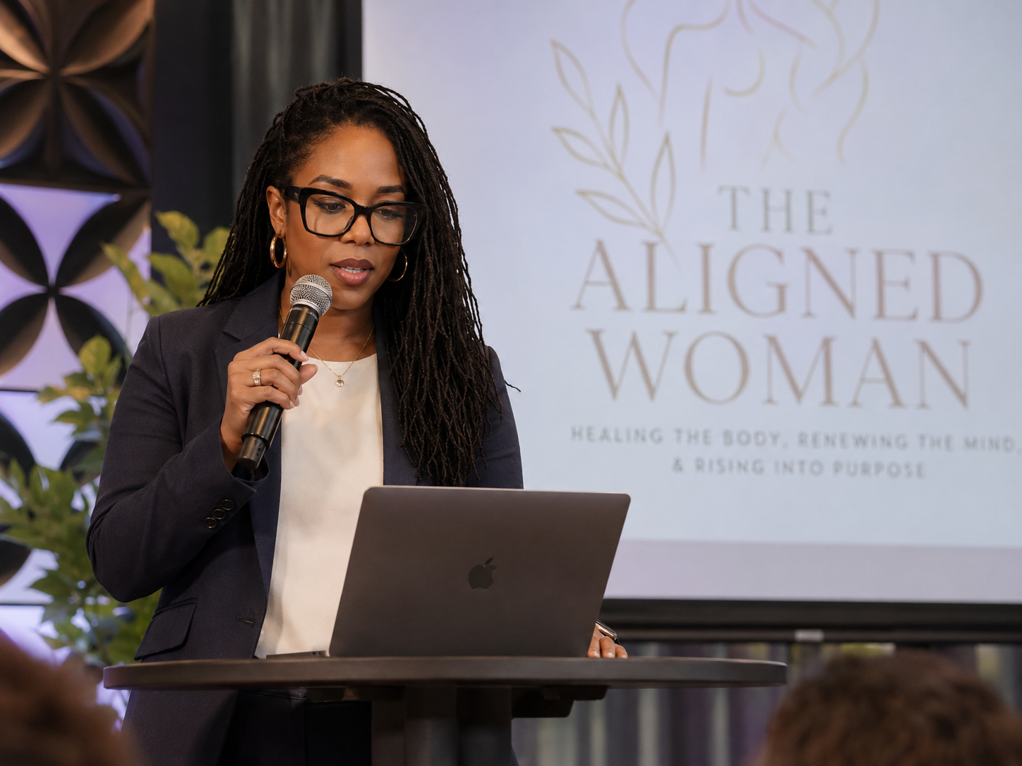 A woman with long dreadlocks, glasses, and gold hoop earrings is speaking into a microphone while standing at a podium with a laptop. Behind her, a screen displays the text "The Aligned Woman" and a subtitle about healing the body, renewing the mind, and rising into purpose.