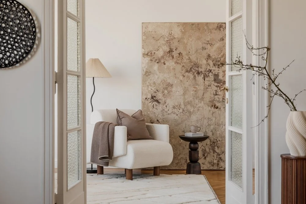 Cozy living room with a white armchair, brown pillow, beige throw blanket, small side table with a cup, textured wall, floor lamp, and decorative branches in a vase.